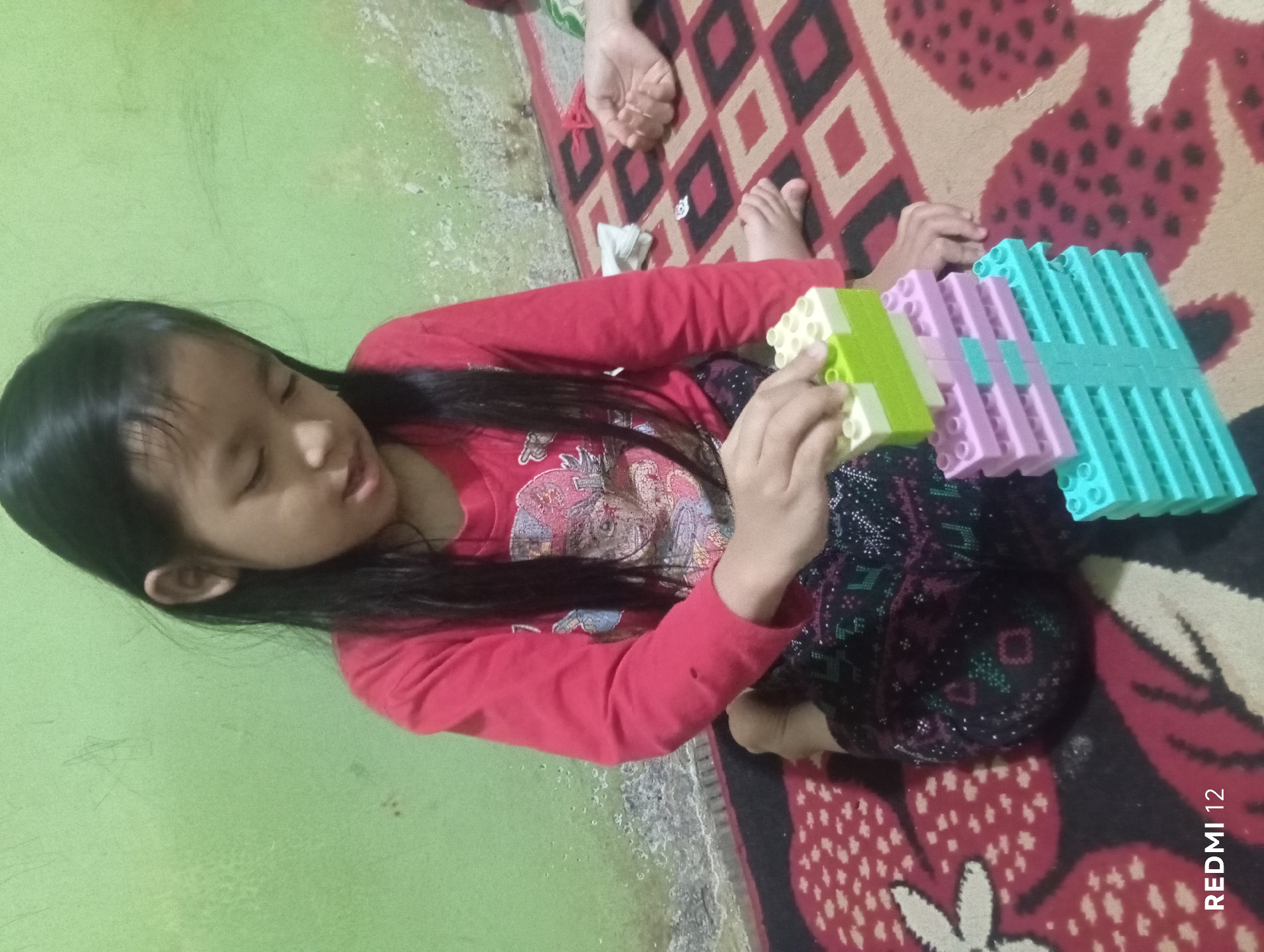 The image shows a young girl with long dark hair wearing a red long-sleeved shirt and patterned pants. She is sitting on a patterned rug and playing with a tower of colorful building blocks. The blocks are stacked in layers of teal, pink, and light green. The girl's hands are visible as she manipulates the blocks. In the background, a light green wall is visible. A second person's hands and feet are partially visible on the rug to the right of the girl. The scene appears to be an indoor setting, likely a home. The image has a watermark at the bottom left corner that reads "REDMI 12". It is not possible to determine the specific location within Pagar Alam, Indonesia from the visual information provided.