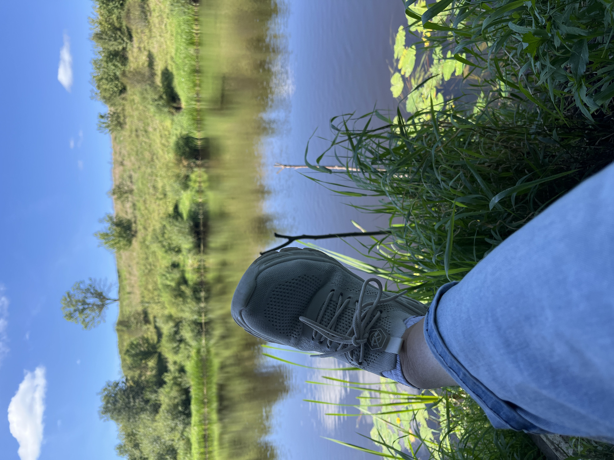 The image depicts a person's leg, clad in light blue denim jeans and a light gray sneaker, resting in the foreground. The leg is positioned such that the sneaker points towards the right side of the frame, with its laces clearly visible. The setting appears to be a natural outdoor environment, likely near a body of water. Tall green grasses and reeds surround the person's leg, with some lily pads floating on the dark water. In the background, a calm river or lake reflects the lush green trees and the blue sky, which is dotted with a few white clouds. The landscape beyond the water is characterized by a gently sloping grassy hill covered in vegetation and scattered trees. The overall scene conveys a sense of tranquility and relaxation in a natural setting. There is no indication of the specific location within Германавіцкі сельскі Савет, Belarus, beyond it being a natural, possibly rural or park-like environment.
