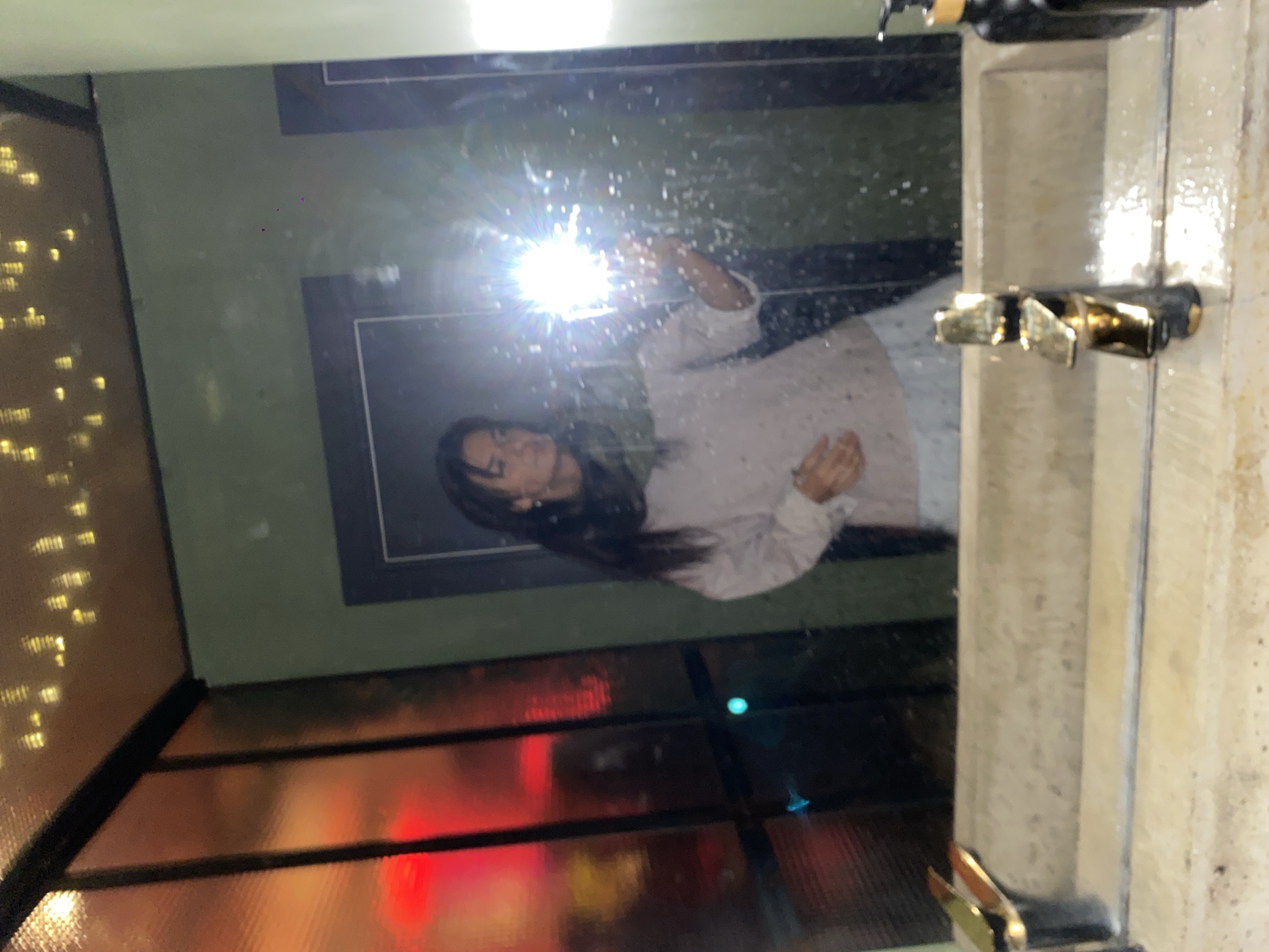 The image depicts a reflection in a bathroom mirror. A woman is visible in the mirror, wearing a light-colored top. The bathroom features a sink with a faucet, and part of a soap dispenser is also visible. The surrounding environment, reflected in the mirror, shows what appears to be interior lighting and possibly a window. The location is identified as Moscow, Russia.