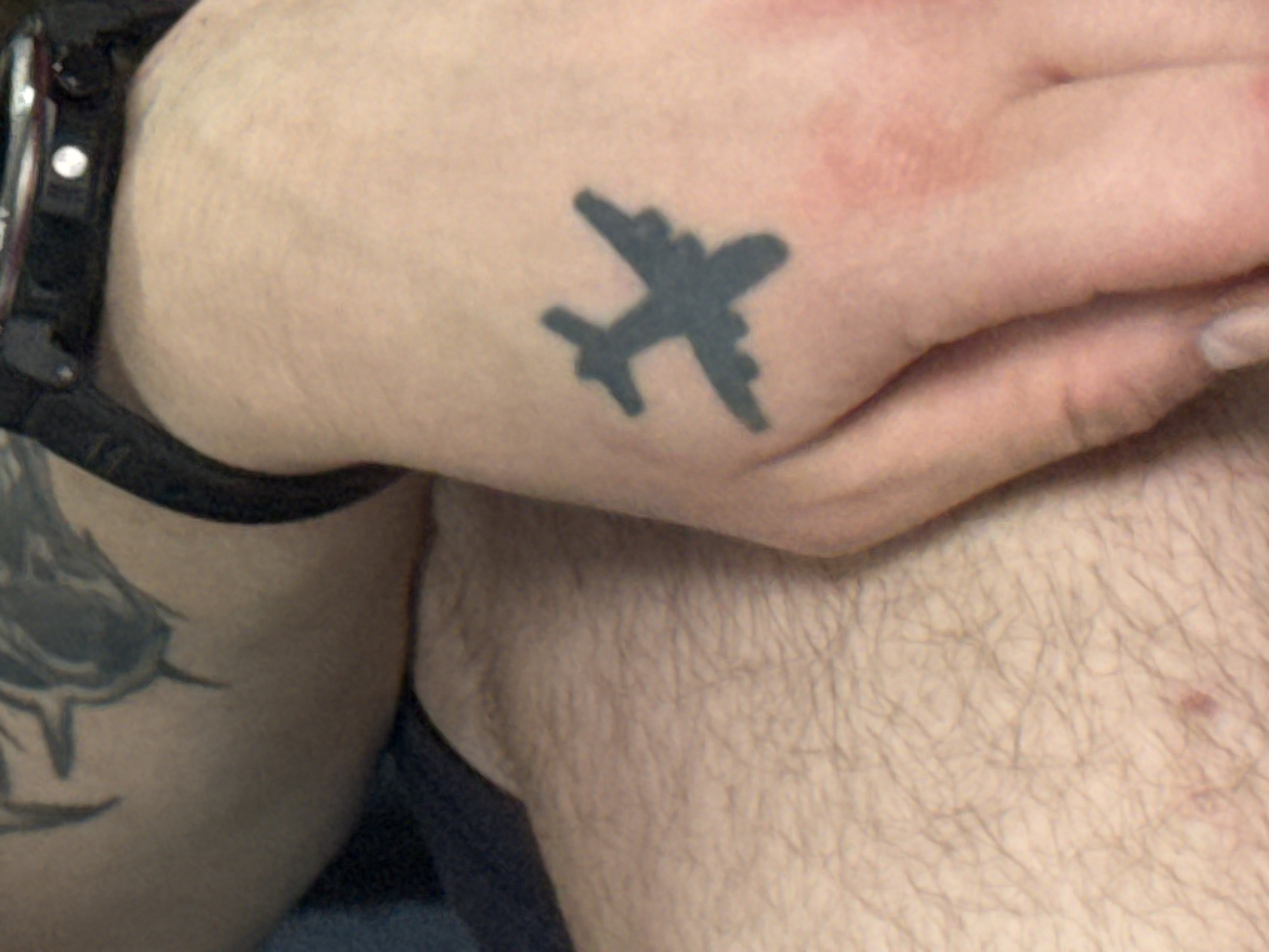 An overhead, close-up shot shows a person's right hand and arm. On the back of the hand, a small, dark tattoo of an airplane is visible. The airplane tattoo is a silhouette with wings and a fuselage. The skin on the hand is light-colored with some faint redness. The person is wearing a dark watch on their left wrist, partially visible at the top of the frame. The background consists of the person's arm, which has a large, faded tattoo of what appears to be a mythological creature, and a portion of their leg covered in light-colored hair. The scene appears to be indoors, with soft, diffuse lighting.
