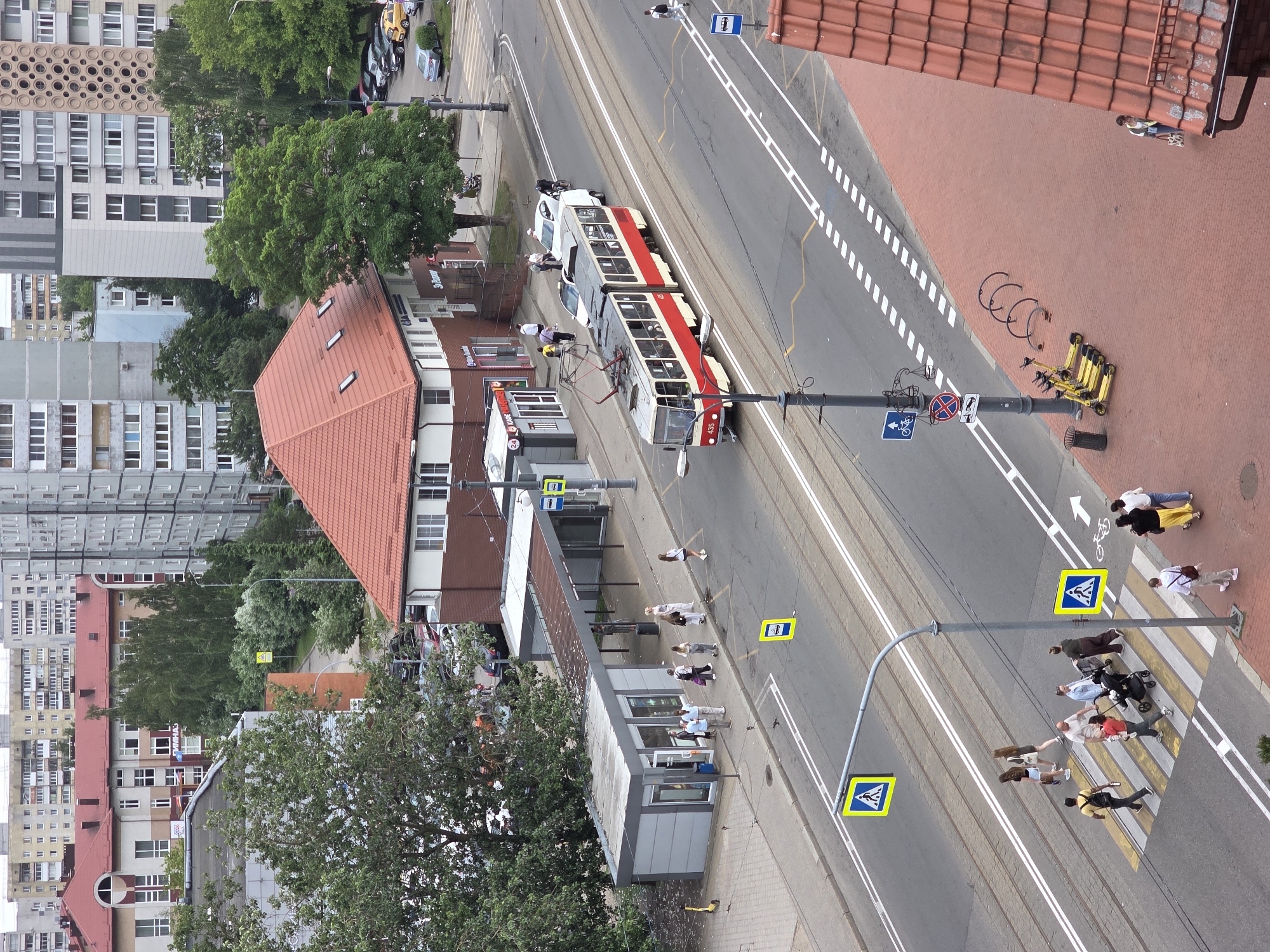 This is an aerial perspective of a bustling urban street scene in Kaliningrad, Russia, observed on an overcast day. In the foreground, the reddish-brown tiled roof of a building is visible on the bottom right. Adjacent to it, a red brick sidewalk features several yellow rental scooters, three bicycle racks, and a dark grey trash bin. A woman with a light-colored top and dark pants walks away from the viewer on this sidewalk. The main focus is a multi-lane asphalt road with double tram tracks running down its center. A large yellow and white striped pedestrian crossing is prominently marked across the street. Numerous pedestrians are utilizing this crosswalk; a group of at least eight individuals includes a person pushing a dark stroller, a woman carrying a small child, and a man with a dark backpack. Other pedestrians walk on the sidewalks flanking the road. On the tram tracks, a two-car tram, predominantly red with white accents and marked "435," is stopped or moving slowly, oriented towards the left side of the frame. Ahead of the tram, a white van and a white car are present on the road, and a cyclist in dark clothing is also visible. Several other vehicles, including a silver sedan and a yellow hatchback, are parked further down the street on the right. To the left of the road, a cluster of modern bus shelters and kiosks is present. One shelter is an enclosed, dark-roofed structure, while another appears to be an open-sided shelter. Several people are gathered near these structures, suggesting a public transport stop. Signage on the street includes multiple yellow diamond-shaped pedestrian crossing signs with a black walking figure, blue rectangular bus stop signs, a blue circular bicycle sign, and a red circular "no stopping" sign. Beyond the immediate street level, various buildings constitute the urban environment. Directly behind the tram stop and bus shelters is a large building with a red tiled roof and light-colored walls, housing businesses on its ground floor with Cyrillic signs such as "ЭКСПРЕСС" (Express) and "КАФЕ" (Cafe). Further into the background, a large, multi-story building with a red roof and prominent arched windows bears a Cyrillic sign that includes "ГИМНАЗ" (Gymnasium). The skyline is dominated by numerous high-rise residential buildings, varying in height and color (greys, beiges, light blue), indicative of a post-Soviet architectural style. One tall building on the right side of the background exhibits