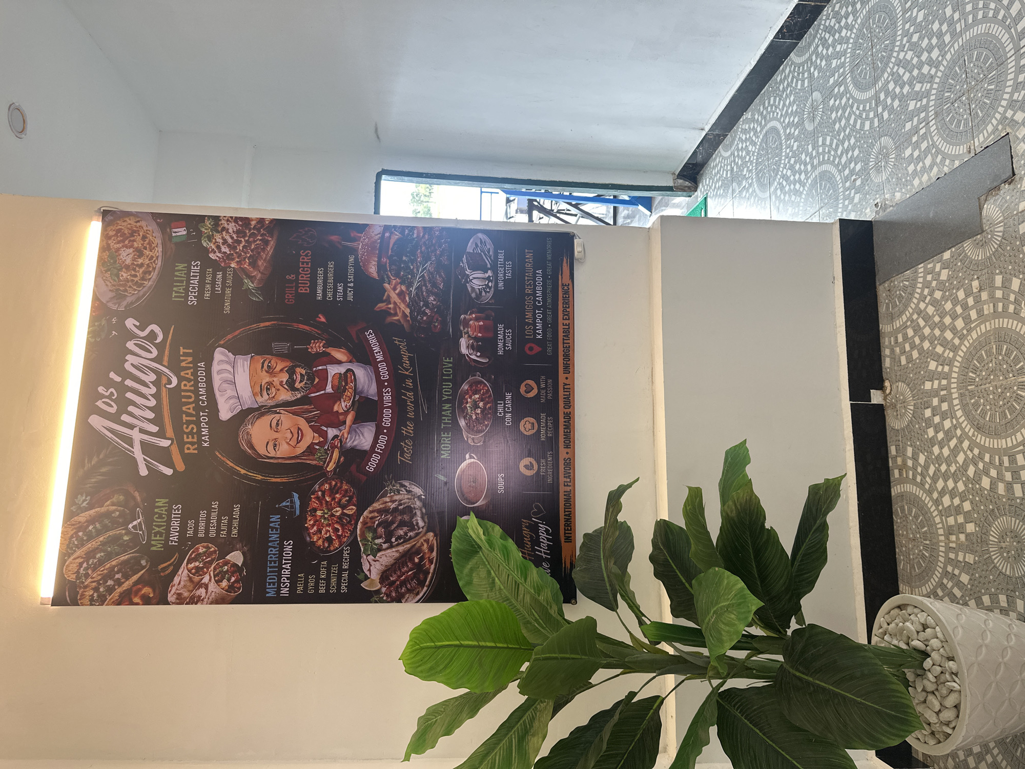 The image shows a large menu poster for "Los Amigos Restaurant" located in Kampot, Cambodia. The poster features various categories of food, including "Mexican Favorites" (tacos, burritos, quesadillas, fajitas, enchiladas), "Mediterranean Inspirations" (paella, gyros, beef kofta, schnitzel, special recipes), "Italian Specialties" (fresh pasta, lasagna, signature sauces), and "Grill & Burgers" (hamburgers, cheeseburgers, steaks, juicy & satisfying). It also highlights "Soups," "Chili con Carne," "Fresh Ingredients," "Homemade Recipes," and "Homemade Sauces." The poster includes caricatures of a male and female chef, along with images of food items. The restaurant's slogan "Hungry We Happy!" and "Taste the world in Kampot!" are prominent. The bottom of the poster states "International Flavors - Homemade Quality - Unforgettable Experience" and provides the full restaurant name and address: "Los Amigos Restaurant, Kampot, Cambodia," followed by "Great Food - Great Atmosphere - Great Memories."
In the foreground, a large green plant in a white decorative pot is visible, partially obscuring the bottom left of the poster. The poster is mounted on a white wall. The floor is tiled with a circular, geometric pattern in shades of grey and white. To the right of the poster, a doorway leads to an outdoor area with tables and chairs visible, suggesting an open-air seating arrangement. The overall environment appears to be an indoor hallway or entrance area of a restaurant.
