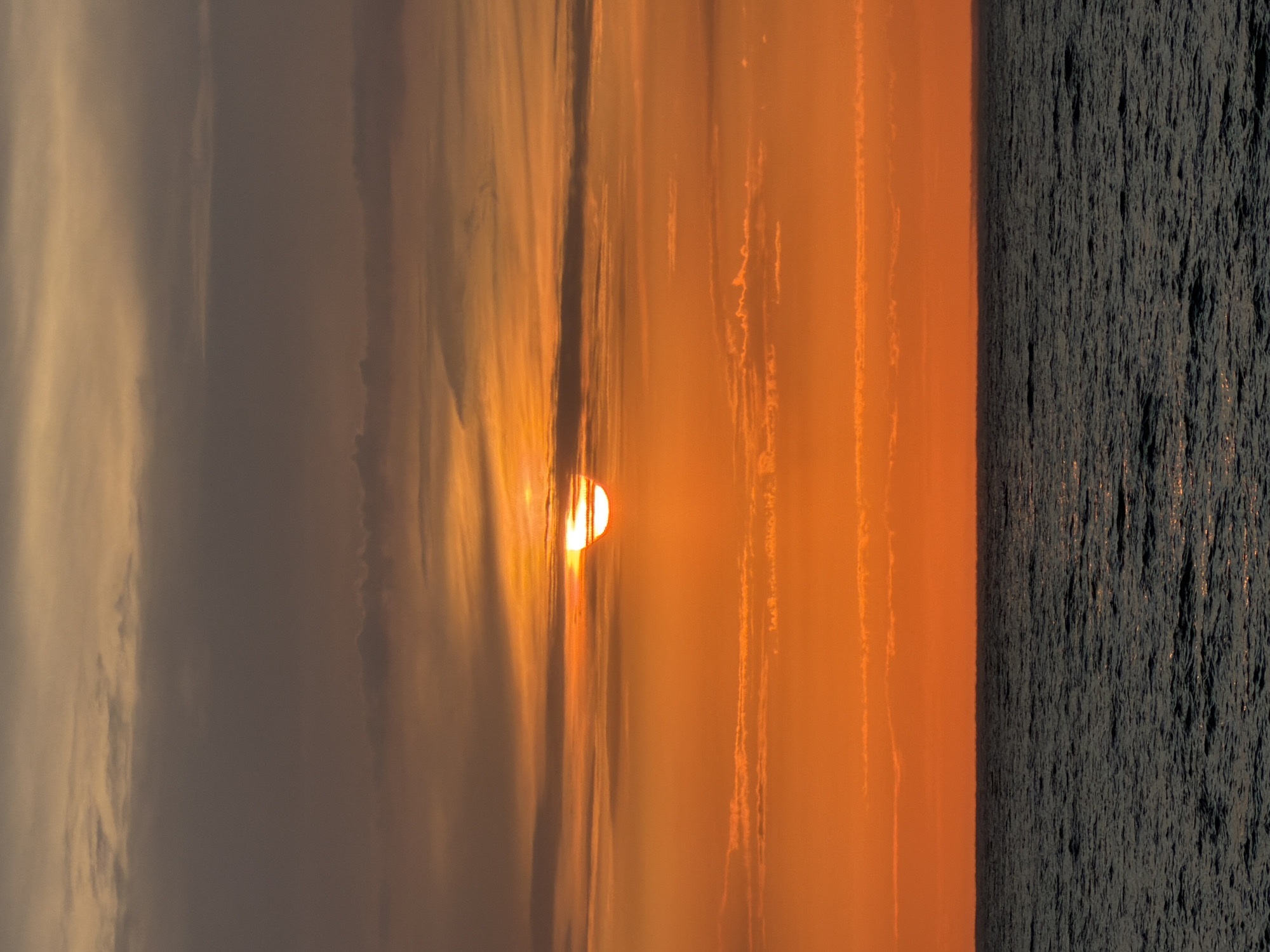 This is a vertical, eye-level, medium shot of a sunset over the Black Sea, with a portion of the sun visible above the horizon. The sky is a vibrant gradient of orange and yellow, with streaks of gray clouds. The sea is dark blue and choppy, with small whitecaps reflecting the light. The scene is peaceful and serene. The location is the coast of Sochi, Russia. There are no people or man-made objects visible in the image.