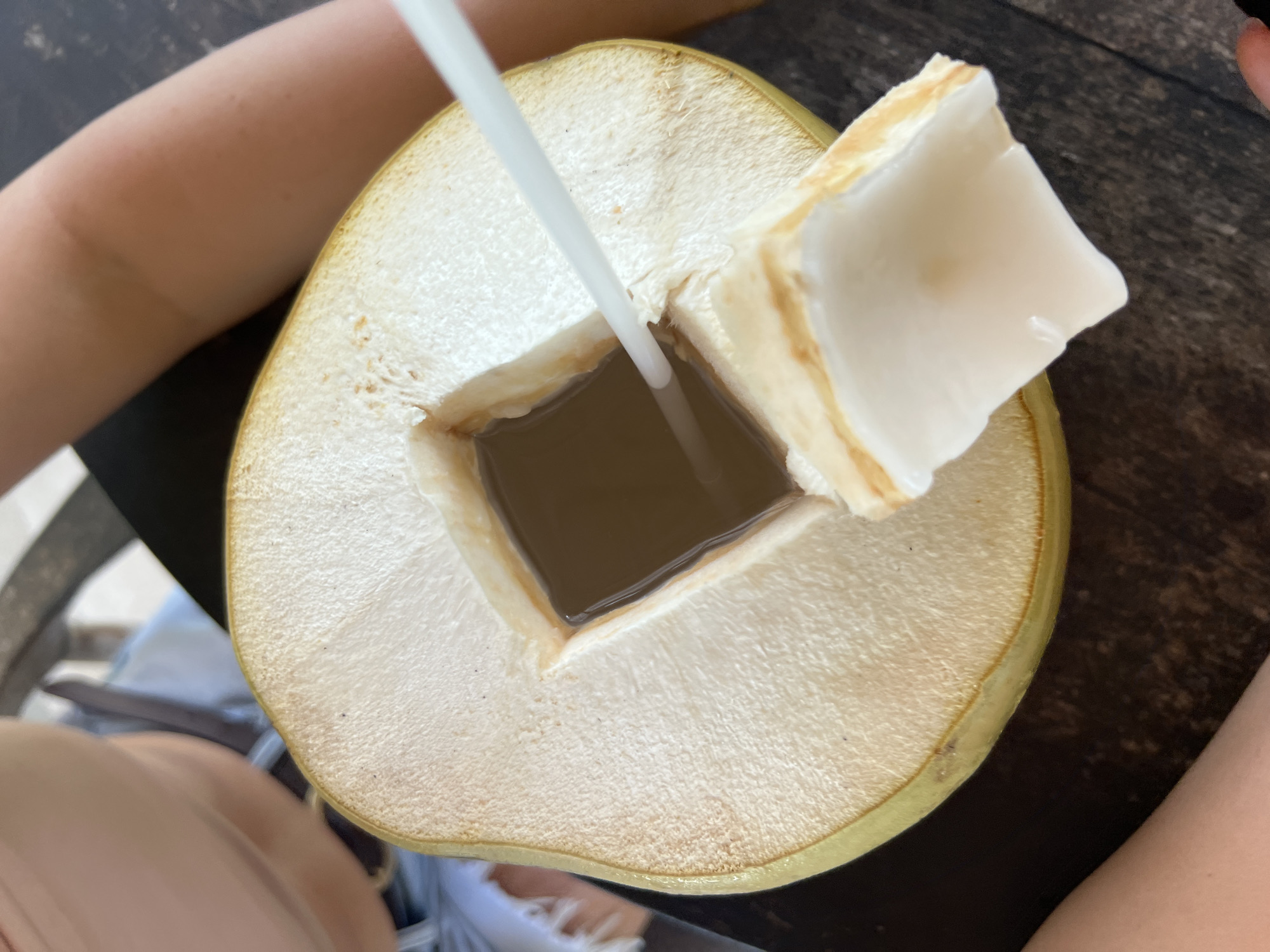 A close-up shot from above captures a young coconut, its top expertly cut open to reveal a square opening with a straw inserted. A rectangular piece of the coconut shell and flesh is folded back, revealing the white flesh. The coconut rests on a dark wooden table. Parts of two people's arms are visible on the left and right sides of the frame, suggesting they are holding or interacting with the coconut. The background is blurred, but hints of clothing and a chair suggest an outdoor setting. While not explicitly stated, the context of the query implies this scene may have occurred in Kuta Selatan, Indonesia, although this cannot be definitively determined from the image.