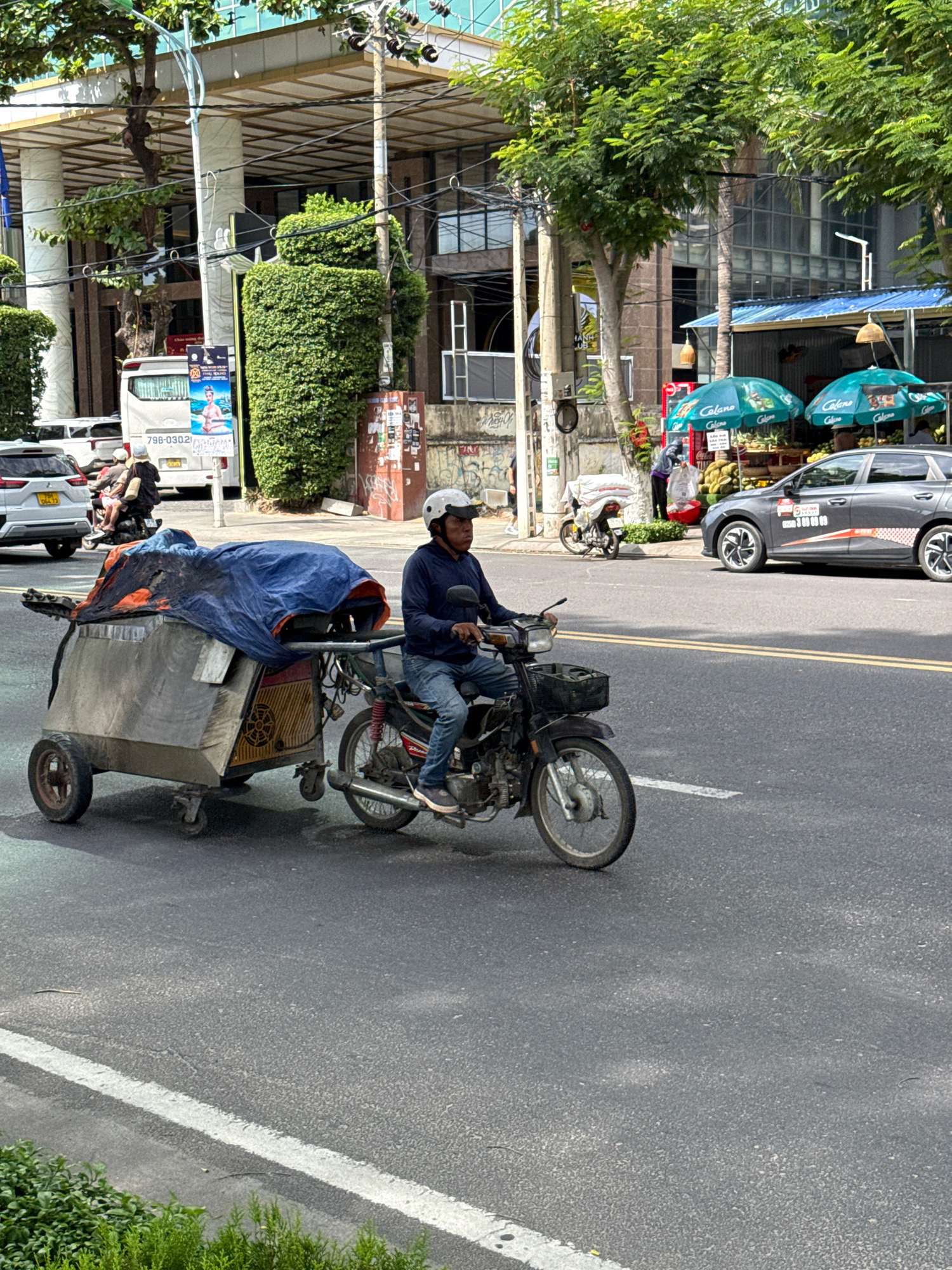 A man rides a motorcycle with a cart attached behind it, on a street in Nha Trang, Vietnam. The man is wearing a blue long-sleeved shirt, blue jeans, and a white helmet. The cart is made of metal and covered with a blue and orange tarp. The motorcycle is black and has a basket in the front. In the background, there are buildings, trees, and other vehicles. The street is paved and has yellow lines marking the lanes. The weather appears to be overcast.