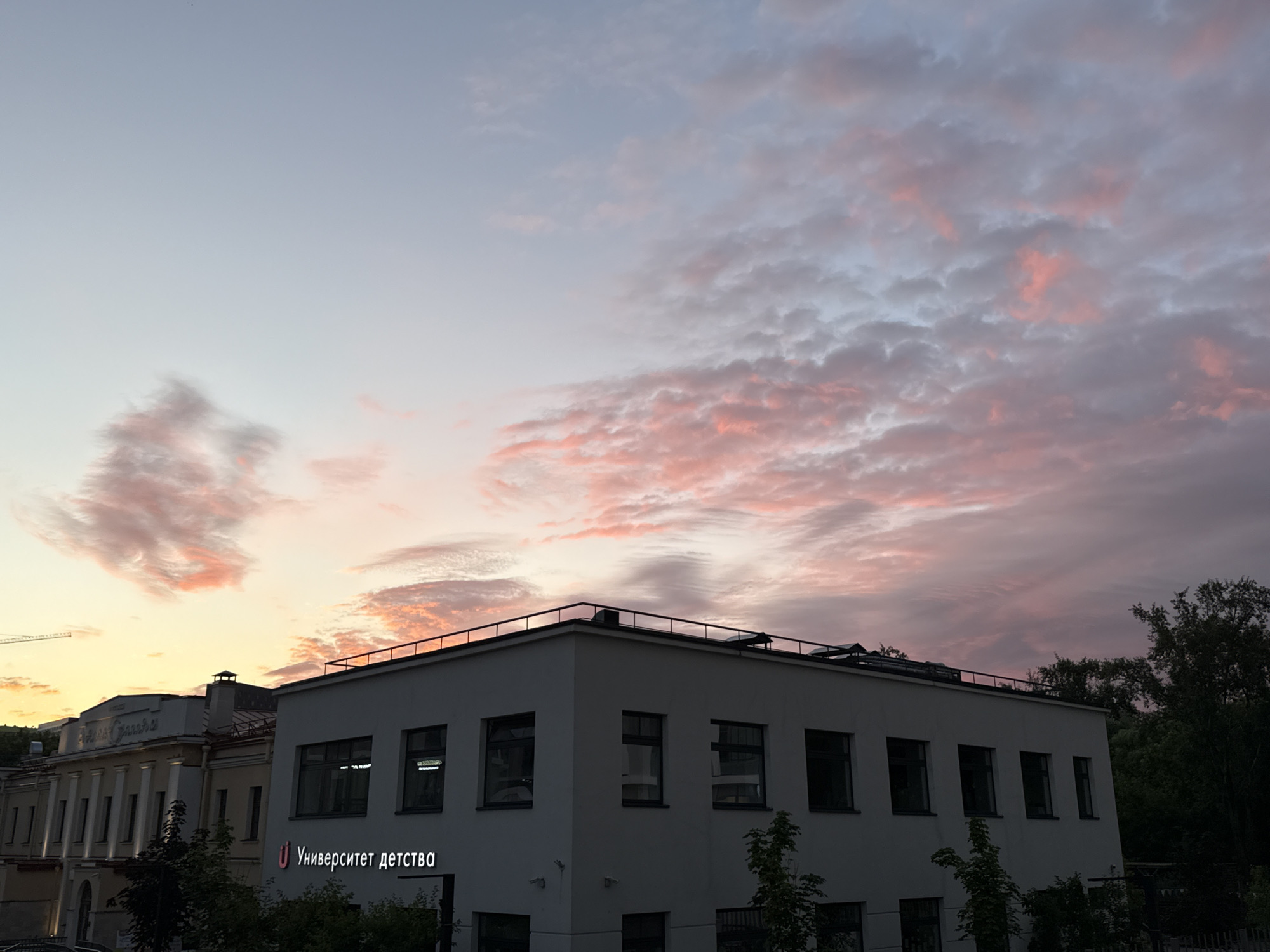 This image captures a twilight sky over Moscow, Russia, with buildings in the foreground. The sky is a gradient of pale blue and pink, illuminated by the setting sun, with scattered clouds that are tinged with vibrant pink and orange hues. In the middle ground, there is a modern, white, rectangular building with dark-framed windows. A sign on this building reads "Университет детства" (University of Childhood). To the left, a more classical-style building with columns is visible, also illuminated by the fading light. Trees frame the right side of the image, their dark silhouettes contrasting with the colorful sky. The overall impression is one of a peaceful urban evening.