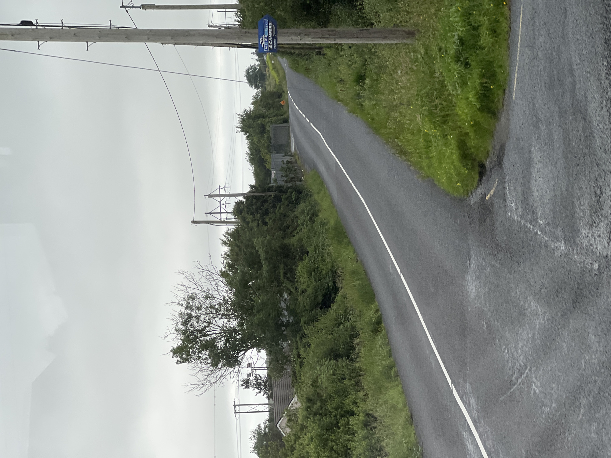 The image depicts a rural road curving to the right in Gweedore, Ireland. The road is paved and has a white line marking its center. On the right side of the road, a wooden utility pole stands tall, with a blue sign attached to it. The sign has white text that reads "CLUB EUROPA & CAMPERS" and "RYELEA". The surrounding environment is lush and green, with grass and trees lining the roadside. In the background, more trees and a house are visible, partially obscured by foliage. The sky is overcast and grey, suggesting a cloudy day. The scene appears to be empty of any people or vehicles, giving a sense of quiet solitude.