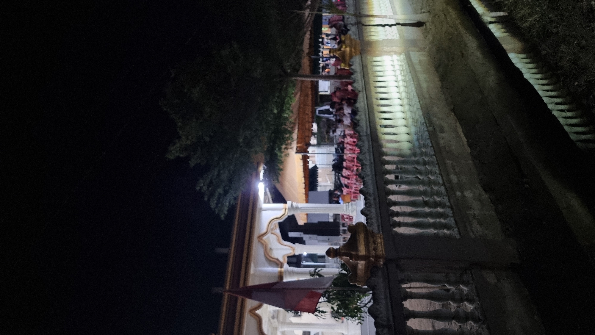 This is a nighttime photograph taken in Manetunong, Indonesia. The scene depicts an outdoor gathering under a canopy of tents, with people seated at tables. In the foreground, there is a decorative white railing with columns, reflected in a body of water. Behind the railing, a building with arched entrances and columns is visible. An Indonesian flag is flying on a flagpole to the left of the building. The overall atmosphere appears to be festive or communal, illuminated by artificial lighting.