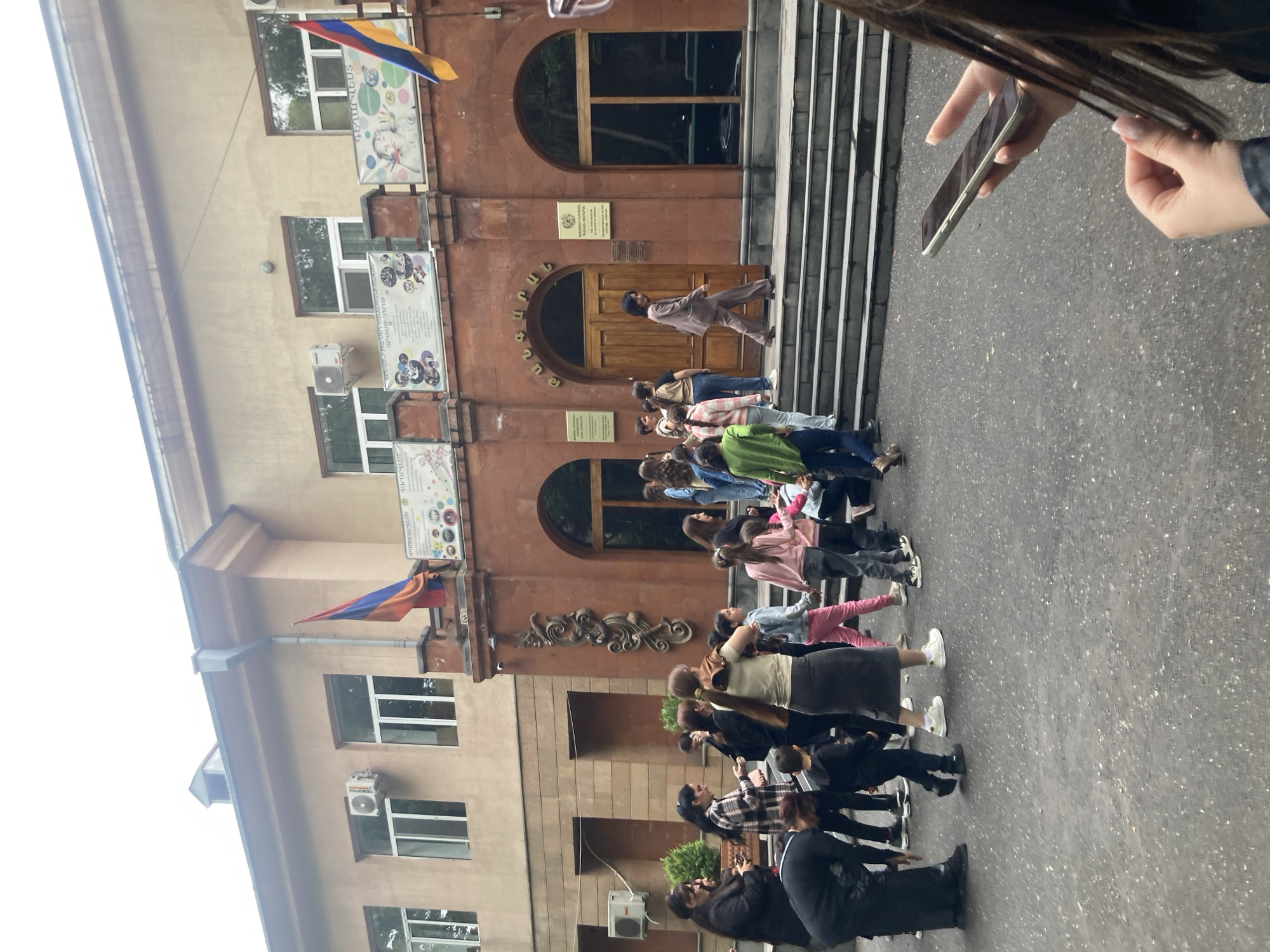 The image depicts a building with the flags of Armenia displayed at the top. A crowd of people are gathered in front of it, some standing on the steps leading to the main entrance. A person is walking up the steps. A hand holds a smartphone at the right side of the frame. Based on the architectural style and the flags displayed, the location may be in Yerevan, Armenia.