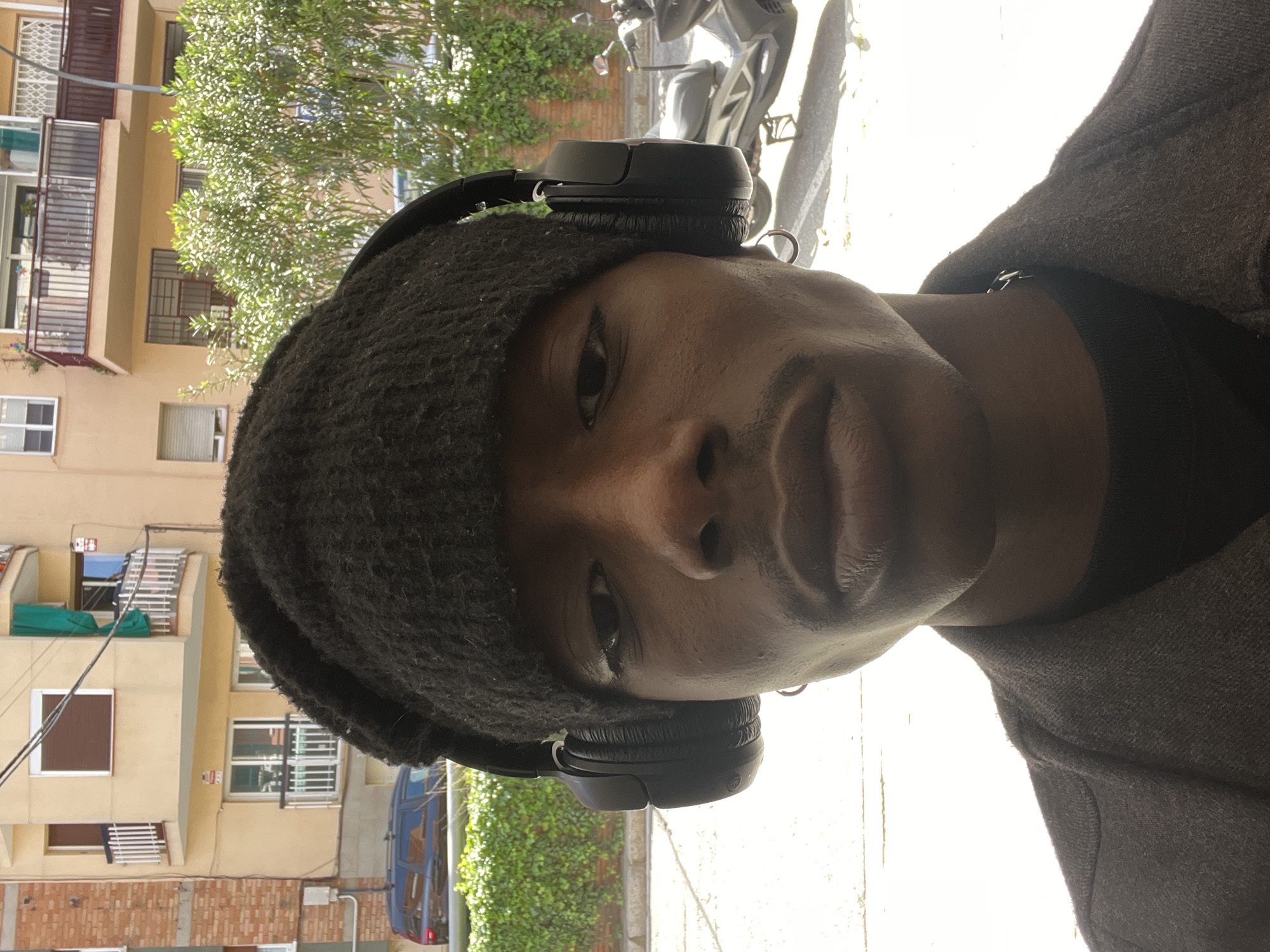 A dark-skinned person wearing a dark beanie and black headphones is shown in a close-up shot. They are wearing a dark shirt and a brown jacket. The location is Barcelona, Spain, evidenced by the architectural style of the apartment buildings in the background. A tree and a parked motor scooter are also visible.