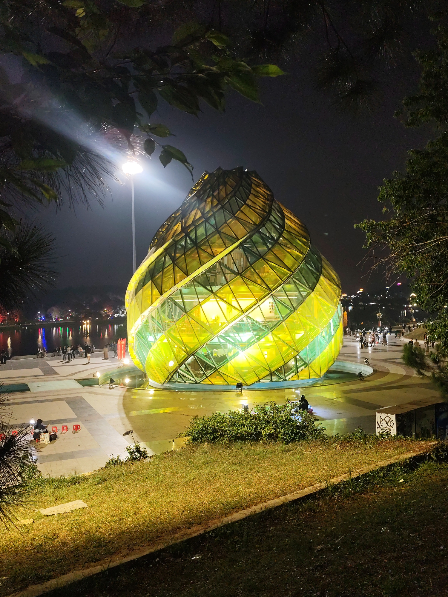 The image depicts Lam Vien Square in Da Lat, Vietnam, at night. The most prominent feature is a large, illuminated glass building resembling a stylized artichoke flower. The structure's glass panels alternate in shades of yellow and green, emitting a bright glow. The building sits on a paved plaza, partially covered with shallow water reflecting the lights.
In the background, across a body of water (presumably Xuan Huong Lake), there are distant city lights. The foreground includes grassy areas and trees framing the scene. People are scattered around the plaza and along the lakefront, suggesting a public space for leisure. A lamppost stands to the left of the artichoke building, casting a strong beam of light. Some graffiti is visible on a concrete structure in the lower right. A few individuals are seated near the edge of the plaza, on red plastic stools.