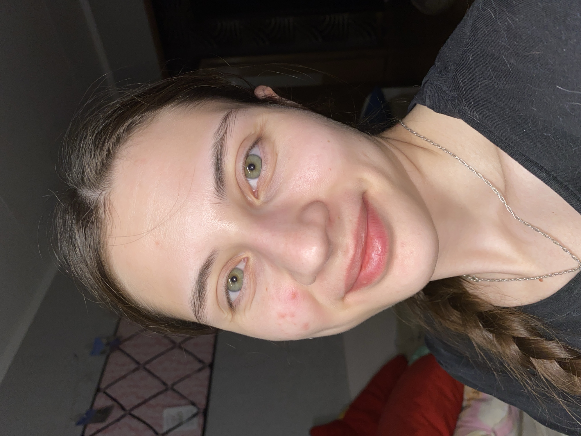 A close-up, angled portrait shows a young woman with fair skin, green eyes, and light brown hair styled in a braid. She is smiling slightly. She is wearing a black top and a delicate silver necklace. Her skin appears clear, with a few small red blemishes on her cheek. The background is indistinct, appearing to show a wall with a grid-like pattern and some furnishings including red pillows. There is no identifiable information in the image to suggest a location in Votkinsk, Russia.