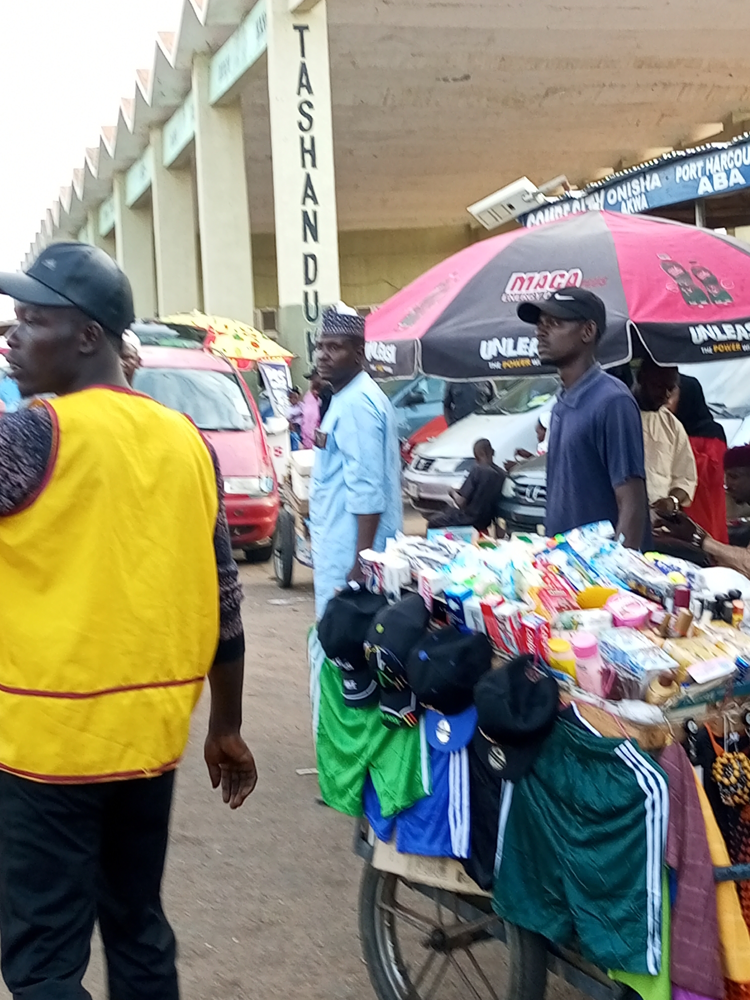 The image depicts a street scene in Gombe, Nigeria, possibly near a market or transportation hub. In the foreground, a man wearing a yellow vest and black pants stands partially to the left, facing away from the camera. To the right of the frame, a vendor's stall displays a variety of items including bottled drinks, toiletries, and packaged goods. Several people are visible in the background, including a man in a blue robe and a traditional cap, and another man in a dark blue t-shirt and a baseball cap. A red vehicle is parked behind the stall, and the scene is framed by a building with distinctive triangular rooflines and prominent signage that reads "TASHANDU". Another sign in the background displays text related to "PORT HARCOURT ONISHA ABA". The overall atmosphere suggests a bustling commercial area.