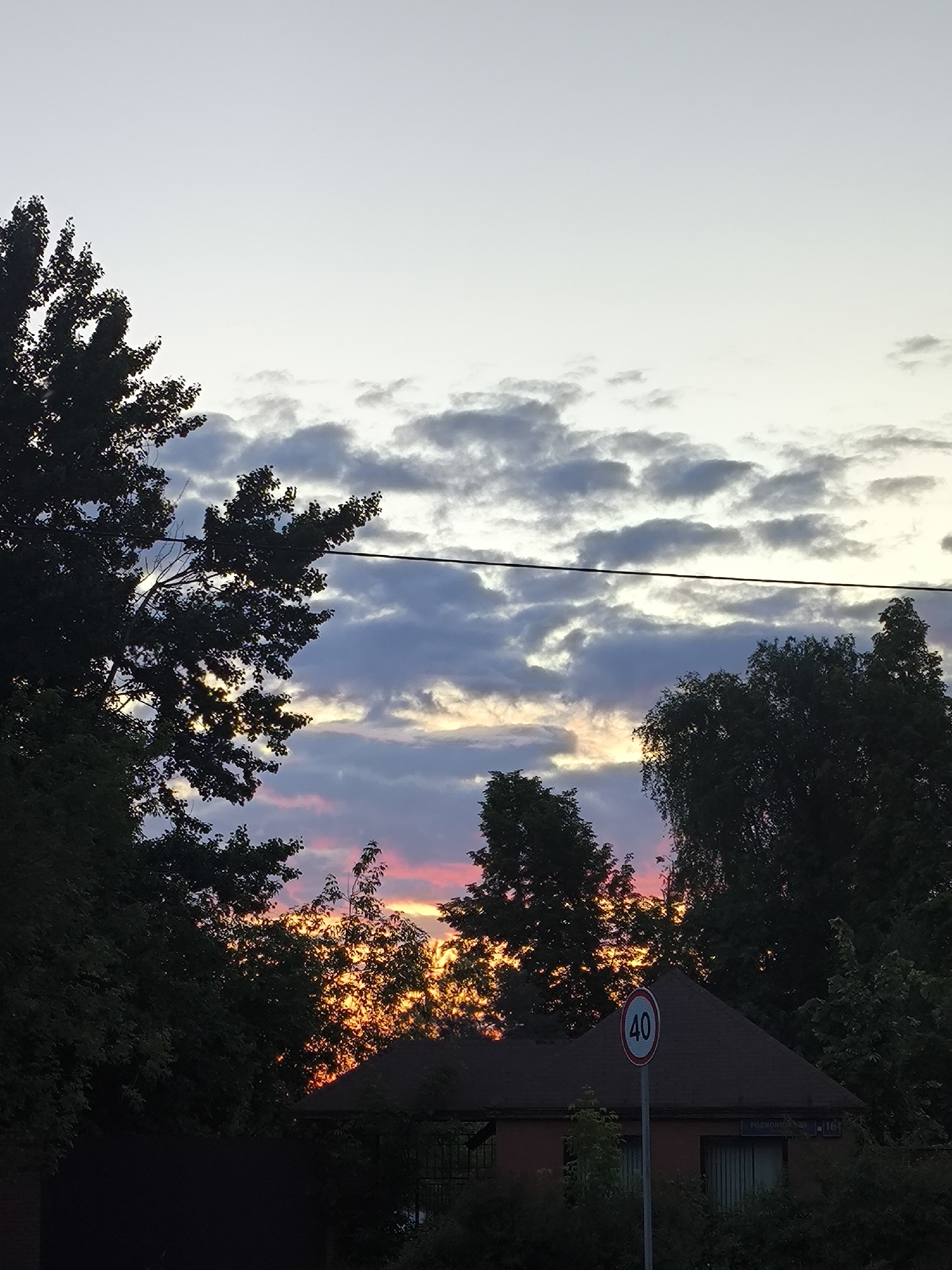 This image captures a vibrant sunset or twilight scene, dominated by a cloudy sky and silhouetted trees. The upper portion of the sky is a pale, light gray, transitioning downwards to patches of darker, somewhat turbulent gray clouds. Beneath these clouds, near the horizon, the sky erupts in brilliant shades of orange and pink, indicative of the sun setting.
In the foreground, numerous trees with dense foliage are silhouetted against the bright sky, framing the colorful horizon. Their dark forms create a strong contrast with the illuminated clouds. A single, thin power line stretches horizontally across the middle of the image, cutting through the sky and partially obscuring some tree branches.
Towards the bottom right, a portion of a house or building is visible, with a dark brown roof and reddish-brown wall. It is largely obscured by the surrounding trees and shrubbery, which are also silhouetted. A circular road sign with a red border and the number "40" in white is prominently displayed on a pole in front of the house, suggesting a speed limit. On the wall of the house, a small rectangular plaque or sign is visible, bearing text in Cyrillic script that appears to read "РОДНОЙ" (Rodnoy) or "РОДНОИ" and "16А".
The overall mood of the image is serene and picturesque, capturing the beauty of a day's end in what appears to be a residential or semi-rural setting. No specific activity is taking place.