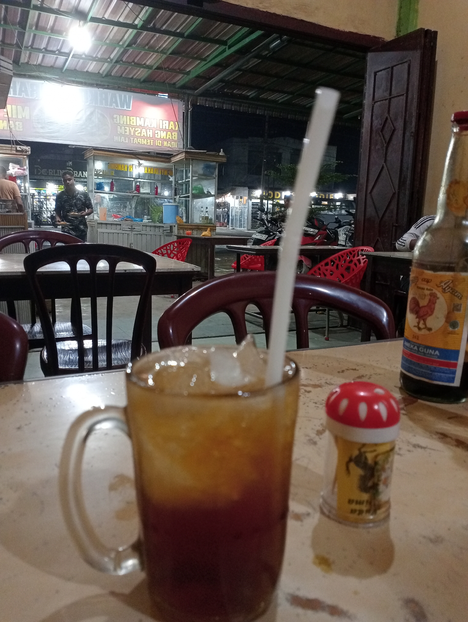 The image, taken in Tingkeummanyang, Indonesia, depicts an outdoor dining scene. A table dominates the foreground, bearing a glass of iced tea with a white straw, a small condiment shaker with a red mushroom-shaped lid, and a bottle labeled "Ayam." The dining area features brown wooden chairs and tables. In the background, illuminated food stalls are visible, with two men standing nearby. Further back, motorbikes are parked. The signage above one stall reads "WARUNG KAMBING," with the words mirrored and inverted in the image.