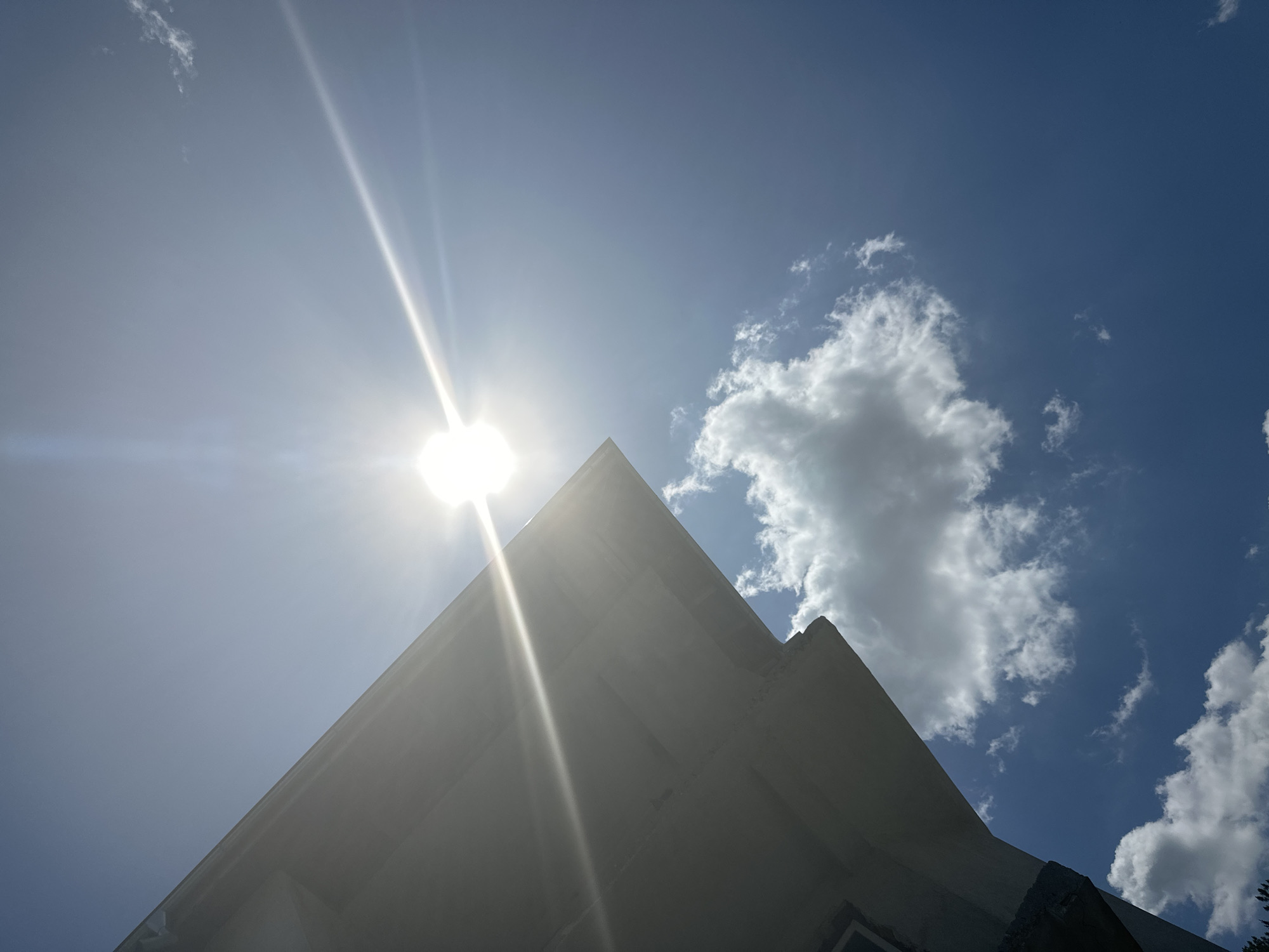 A low-angle shot captures a bright sun with lens flare against a clear blue sky. The edge of a building's eave, likely a house, juts into the frame from the left. Several fluffy white clouds are scattered across the sky, with one larger cloud positioned below and to the right of the sun. The scene appears to be outdoors on a sunny day.