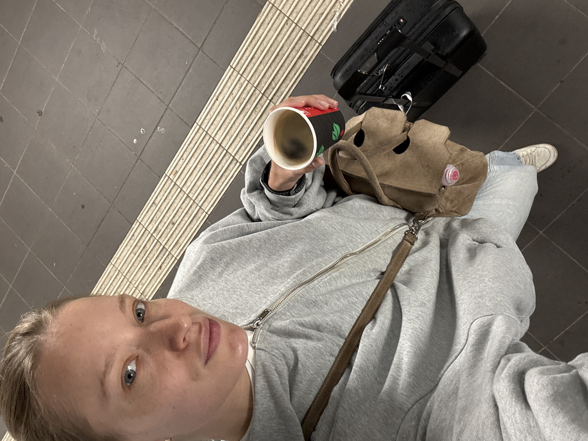A young woman with blonde hair pulled back stands on a train platform in Oelde, Germany, holding a cup of coffee and a brown messenger bag. She is wearing a grey zip-up hoodie, light blue jeans, and white sneakers. To her right, a black suitcase rests on the platform. The platform has a ribbed safety strip along the edge. The woman is looking towards the camera, with a neutral expression.