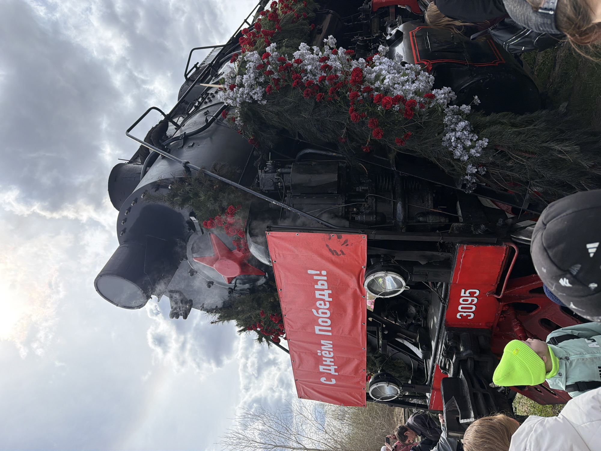 The image captures a decorated steam locomotive, identified by the number 3095 on its front, during a daytime event in Votkinsk, Russia. The train's front is elaborately adorned with abundant red and white artificial flowers, resembling carnations, and intertwined with green pine or fir boughs, suggesting a festive or commemorative occasion. A prominent red banner hangs from the front of the locomotive, displaying the Russian text "С днём Победы!", which translates to "Happy Victory Day!". A red star emblem is also visible on the front of the train.
A crowd of people, including many children, are gathered around the locomotive, looking at it with interest. Several individuals are partially visible in the foreground, with one child wearing a bright neon yellow beanie. The sky above is overcast and filled with light gray clouds, indicating a cloudy day. Steam appears to be emanating from the locomotive, suggesting it is either active or recently active. The overall scene conveys a celebratory atmosphere, likely commemorating Victory Day.
