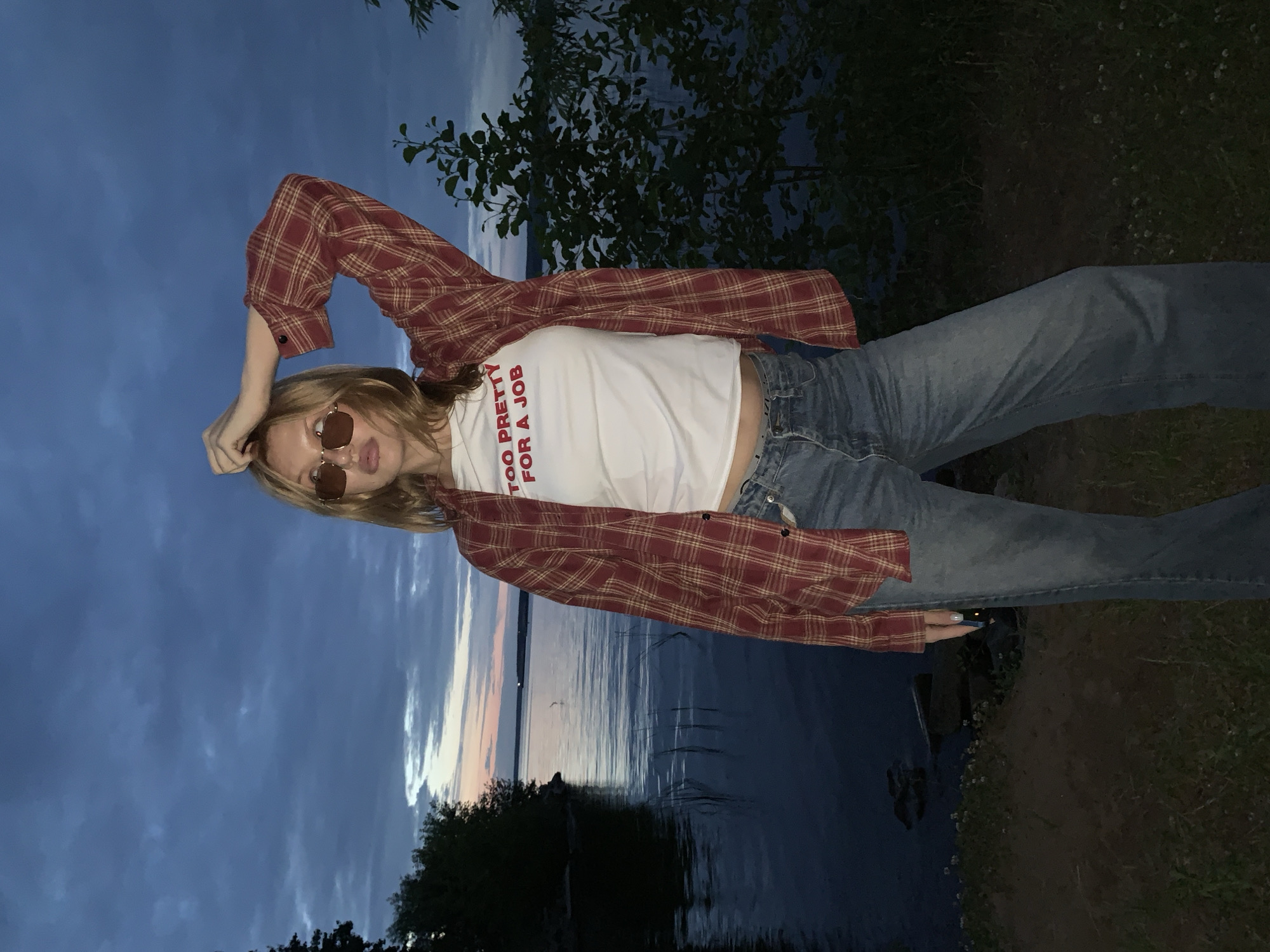 A young woman with blonde hair and sunglasses stands near a body of water at dusk. She is wearing a red and yellow plaid open shirt over a white t-shirt with the text "TOO PRETTY FOR A JOB" in red letters, and blue jeans. She has her right hand raised to the back of her head. The background shows a dark sky with clouds, a calm body of water with ripples, and dark trees on the shore. The location is the Kama River embankment in Chaikovsky, Russia.