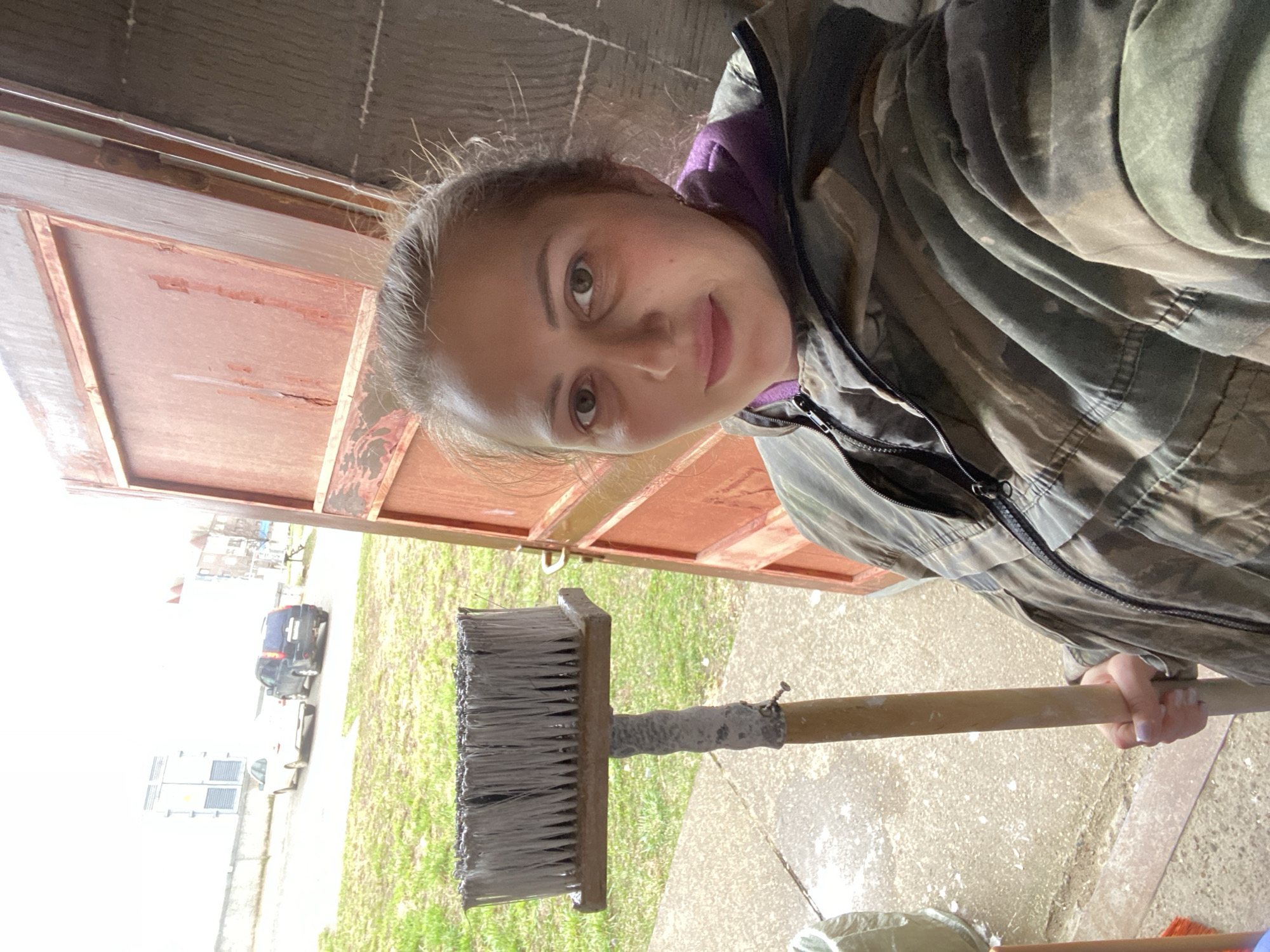 A young woman is depicted in a selfie-style photograph, holding a large paint or whitewash brush outdoors, likely in the doorway of a garage or utility building. She appears to be engaged in a painting or coating project, as the broad, rectangular brush and its wooden handle are heavily covered in a white substance.
The woman has light skin, dark eyes, and light brown or blonde hair pulled back. She is wearing a purple hooded top under a camouflage-patterned jacket or utility coat, partially unzipped. Her gaze is directed at the camera with a calm, neutral, or slightly smiling expression.
The immediate setting includes a concrete ground with white splatters, consistent with a work area. Behind her, a robust red or orange metal door frame, possibly for a garage, is visible. The background opens to an outdoor environment under bright, diffused daylight, suggesting an overcast sky or bright conditions. A paved road leads away from the structure, with a dark-colored car and a lighter vehicle parked along its edge. Further in the distance, multi-story buildings and what appear to be utility boxes or structures can be seen, with patches of green grass in between. The scene takes place in what appears to be a semi-urban or residential area in Марковский, Russia. No visible text is present in the image.