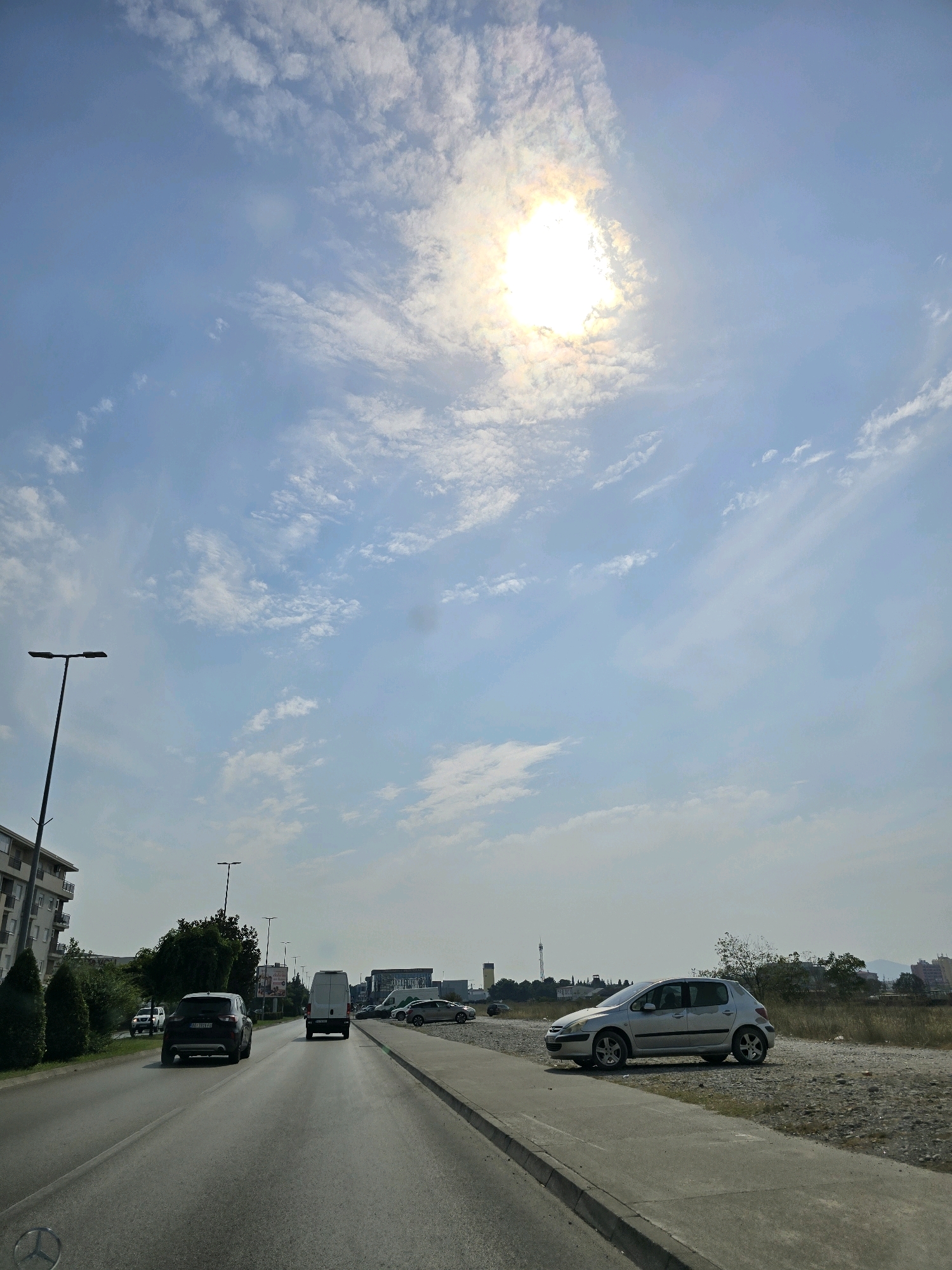 The image shows a view from a vehicle driving on a multi-lane road in the city of Podgorica, Montenegro. The road curves to the right and has clear lane markings. On the left side of the road, there is a sidewalk, a row of trees, and a multi-story building. On the right side of the road, there is a gravel area with parked cars, including a silver Peugeot hatchback and a white van. The sky is blue with scattered clouds, and the sun is visible, partially obscured by clouds, casting a bright light. In the distance, there are buildings and some industrial structures. The overall impression is of a sunny day in an urban or suburban environment.