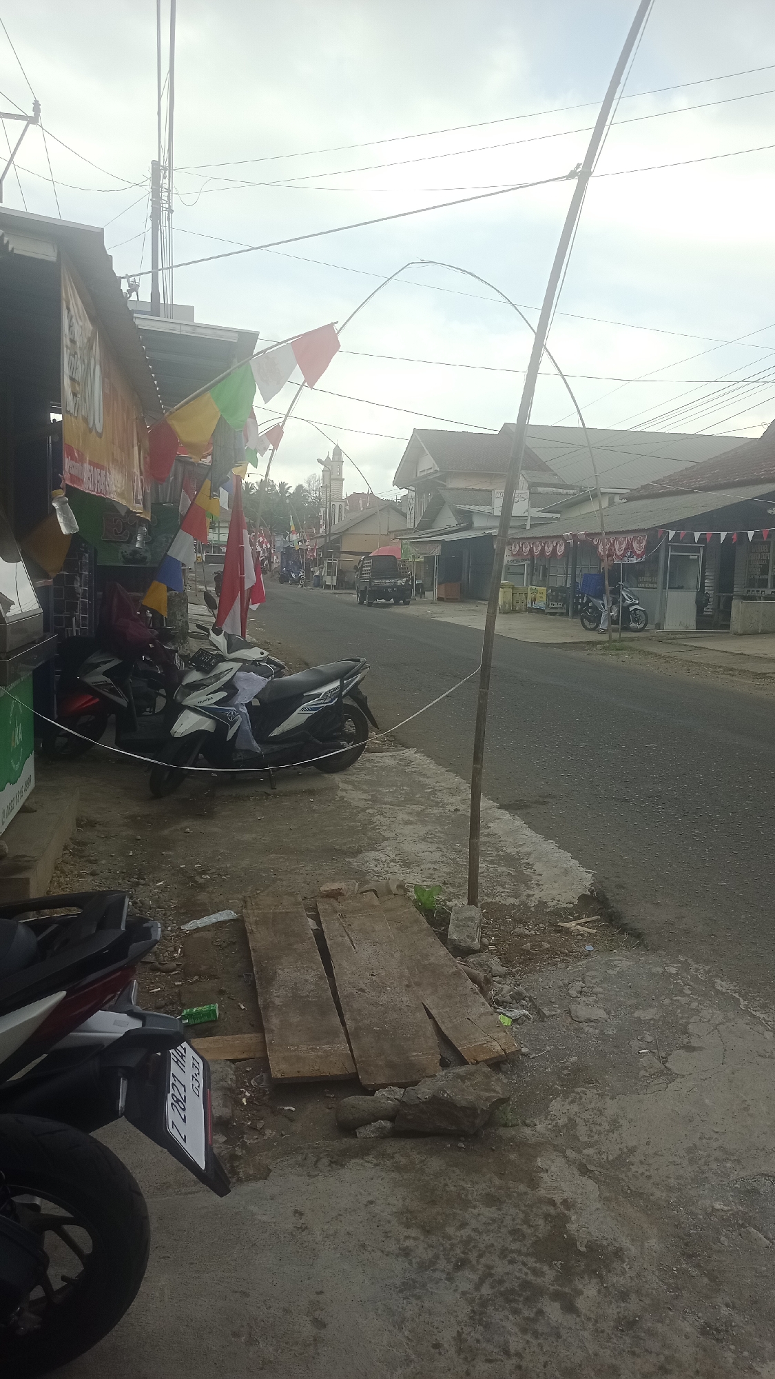 The image depicts a street scene in Cikalong, Indonesia, on an overcast day. A paved road runs through the center of the frame, bordered by a dirt and concrete sidewalk on the right and a row of buildings on the left.
On the left side of the road, several buildings appear to be small shops or businesses. One building has a corrugated metal roof and displays banners and flags, including red and white Indonesian flags and multi-colored triangular flags, suggesting a festive or commemorative occasion. Several motorcycles are parked in front of these establishments, including a white and black scooter and a red and black scooter.
The right side of the road shows a more residential or commercial area with buildings featuring sloping roofs. A tall wooden pole stands on the sidewalk, strung with wires and a string of flags that arc across the street. Further down the road, a pickup truck is visible.
The foreground of the image features a concrete sidewalk with several pieces of rough lumber laid out. Rocks and some scattered debris are also present on the ground. The overall impression is a typical Indonesian street scene, possibly decorated for a local event.