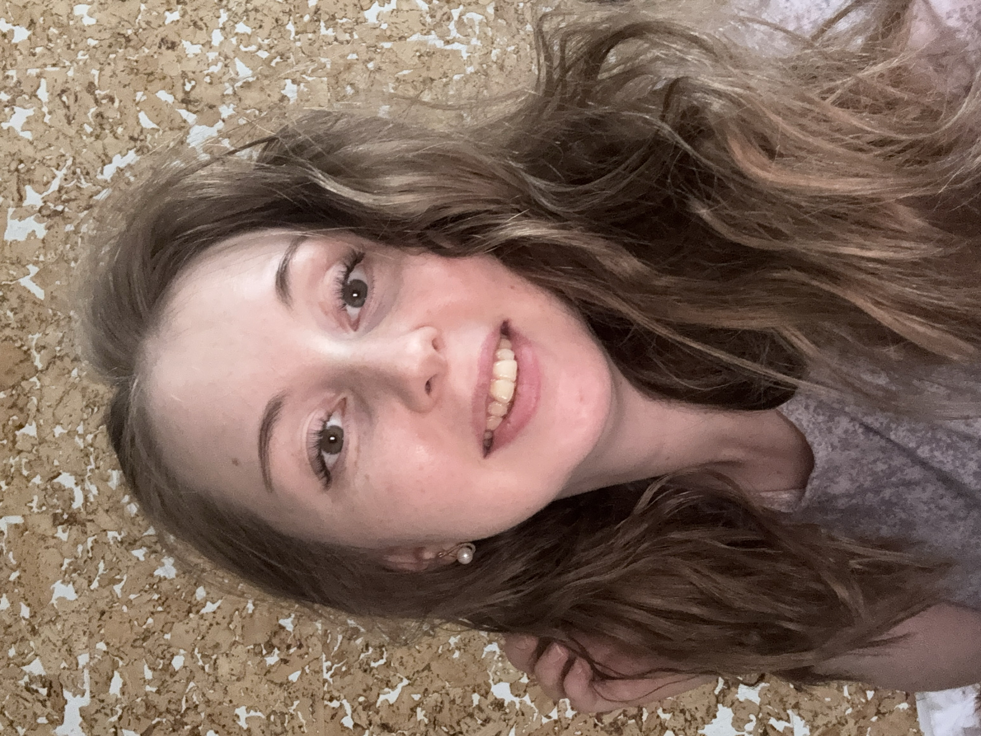 A young woman with long, wavy brown hair is smiling at the camera. She is wearing a light-colored top and a pearl earring. The background is a textured, cork-like surface. The location appears to be indoors.