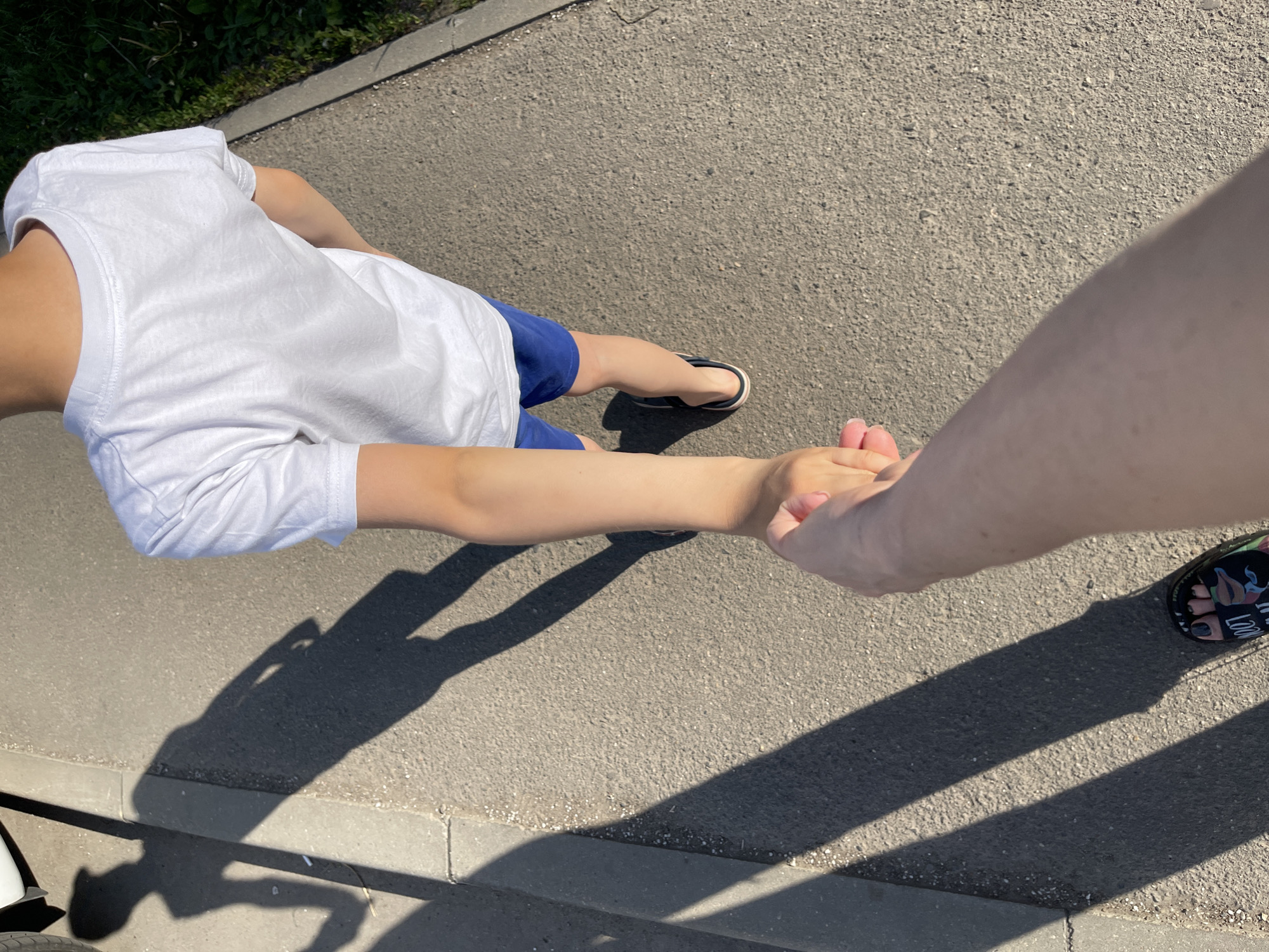 An elevated, first-person perspective captures two individuals walking on an asphalt pathway under direct sunlight. The camera holder's right arm, hand, and partial right leg with a black sandal (featuring patterned straps and dark-painted toenails) are visible at the bottom of the frame. The camera holder's hand grips the right hand of a second individual, a child, who is seen from behind, walking ahead. The child wears a plain white short-sleeved t-shirt, blue shorts, and dark-colored flip-flops with a light trim.
The pathway is a light gray asphalt surface. To the left, a concrete curb separates the pathway from an adjacent area, where a portion of a white vehicle bumper is visible in the top left corner. To the right, a narrow strip of concrete borders a patch of green vegetation. Long, distinct shadows of both individuals are cast on the asphalt, extending diagonally to the left, indicating the sun's position. The scene is located in Kommunarka, Russia.