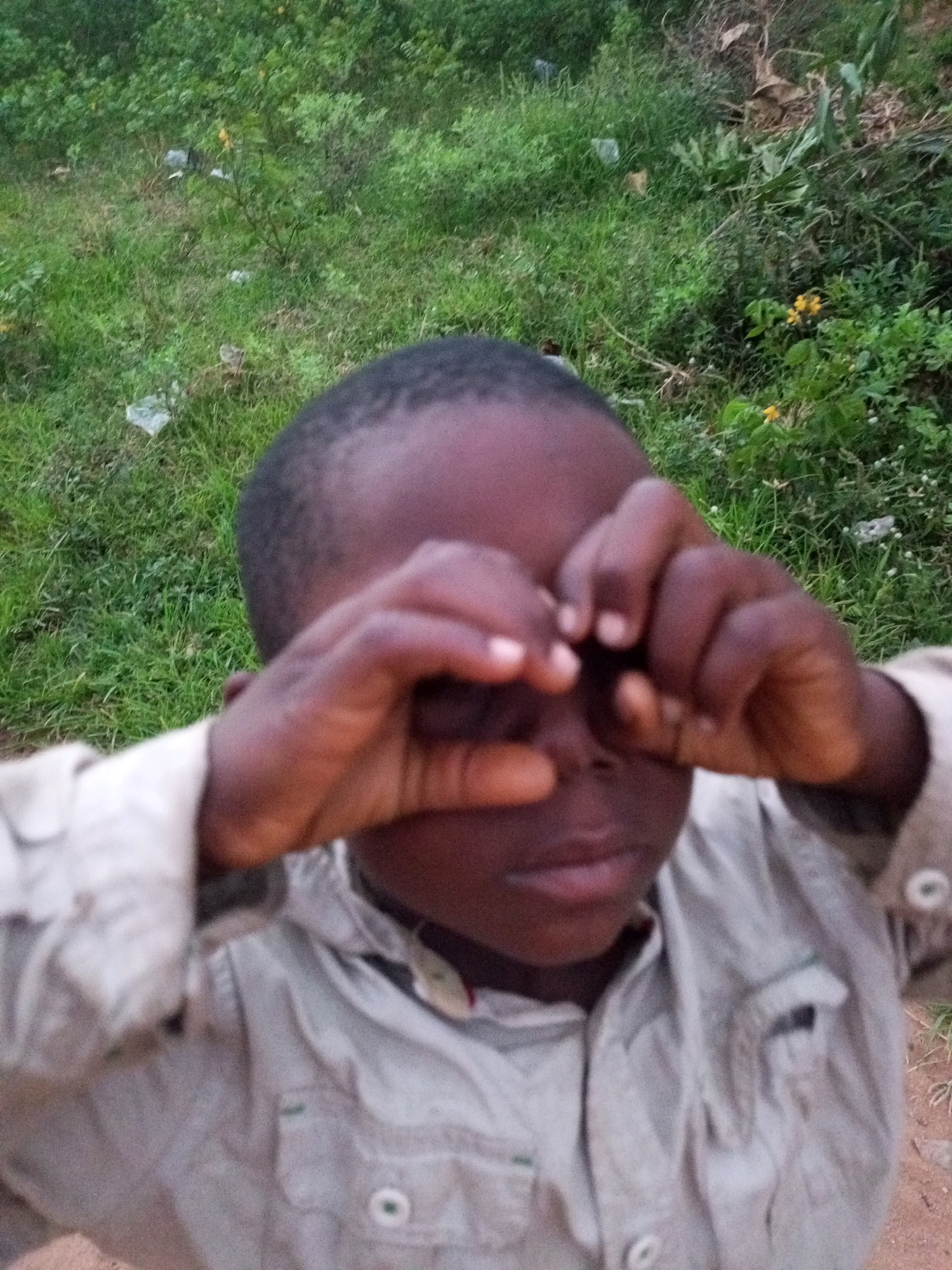 A young child, appearing to be of African descent, is shown from the chest up, looking directly at the camera. The child has dark skin and short, dark hair, and is wearing a light-colored collared shirt with buttons. The child's hands are raised to their face, with fingers near their eyes, as if peering through them. The background consists of green grass and foliage, with some scattered debris, suggesting an outdoor setting, possibly in a rural area or park, within the city of Tempure, Nigeria. The lighting is natural, indicating it is daytime.