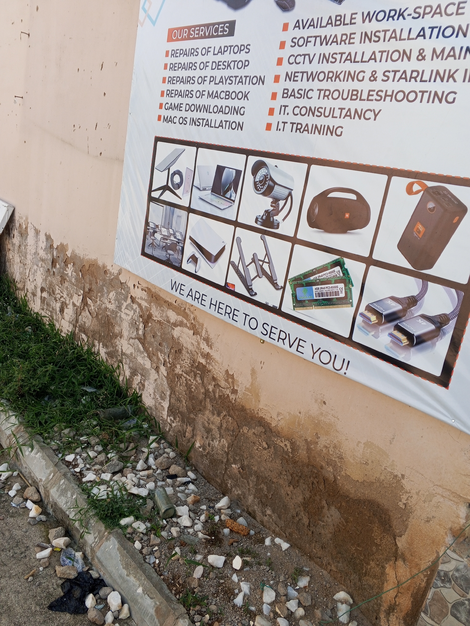 The image shows a faded banner attached to a damaged, weathered wall. The banner advertises "OUR SERVICES" which include "REPAIRS OF LAPTOPS," "REPAIRS OF DESKTOPS," "REPAIRS OF PLAYSTATION," "REPAIRS OF MACBOOK," "GAME DOWNLOADING," "MAC OS INSTALLATION," "AVAILABLE WORK-SPACE," "SOFTWARE INSTALLATION," "CCTV INSTALLATION & MAINTENANCE," "NETWORKING & STARLINK INSTALLATION," "BASIC TROUBLESHOOTING," "IT. CONSULTANCY," and "I.T TRAINING." Below the text are various images of computer hardware and accessories. The wall itself is peeling and discolored, with patches of dampness. In the foreground, a concrete curb runs along the edge of a sidewalk, littered with rocks, debris, and some green grass growing through the cracks. The environment appears to be an outdoor urban setting, likely in Jalingo, Nigeria, given the context. There are no people visible in the image.