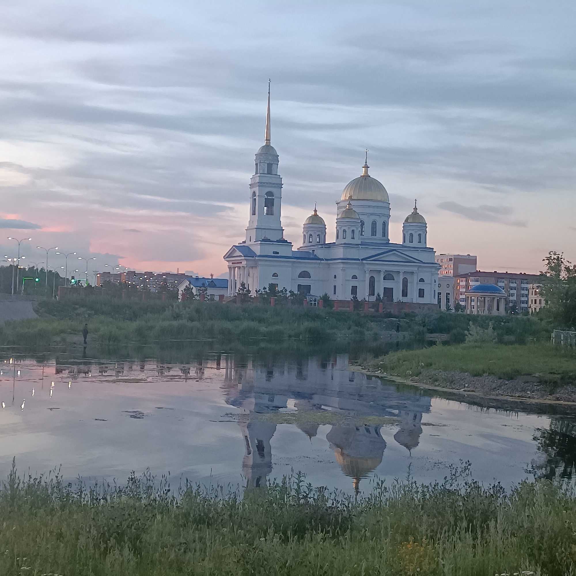 The image depicts a grand white cathedral with golden domes, reflected in a body of water in the foreground. A tall bell tower with a spire stands to the left of the main building. The cathedral is situated on a slightly elevated grassy embankment, with trees and shrubs bordering it. In the background, beyond the cathedral, are several multi-story buildings and streetlights, suggesting an urban environment. The sky is overcast with soft pink and grey clouds, indicative of either dawn or dusk. In the water, the reflection of the cathedral and the sky is clearly visible, creating a symmetrical effect. A single person is visible standing near the water's edge on the left side of the embankment, appearing to be fishing. The overall scene is serene, capturing the architecture of the cathedral and its reflection against a tranquil sky and calm water. The specific location of this cathedral in Kokshetau, Kazakhstan, is the Church of the Intercession of the Holy Mother of God, also known as the Kokshetau Orthodox Cathedral.