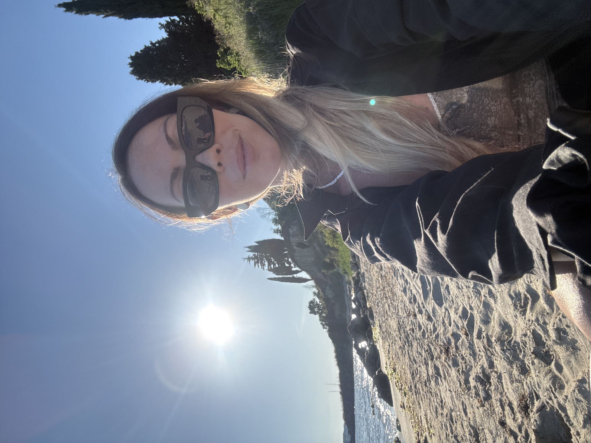 A woman with blonde hair, wearing sunglasses and a black jacket over a patterned top, is taking a selfie on a sandy beach in Nesebar, Bulgaria. The sun is shining brightly in a clear blue sky, casting a glare on the water to her left. The beach is composed of sand and scattered rocks. In the background, there is a gentle slope covered with greenery and trees, including several tall, slender cypress trees. The sea is visible with small waves.
