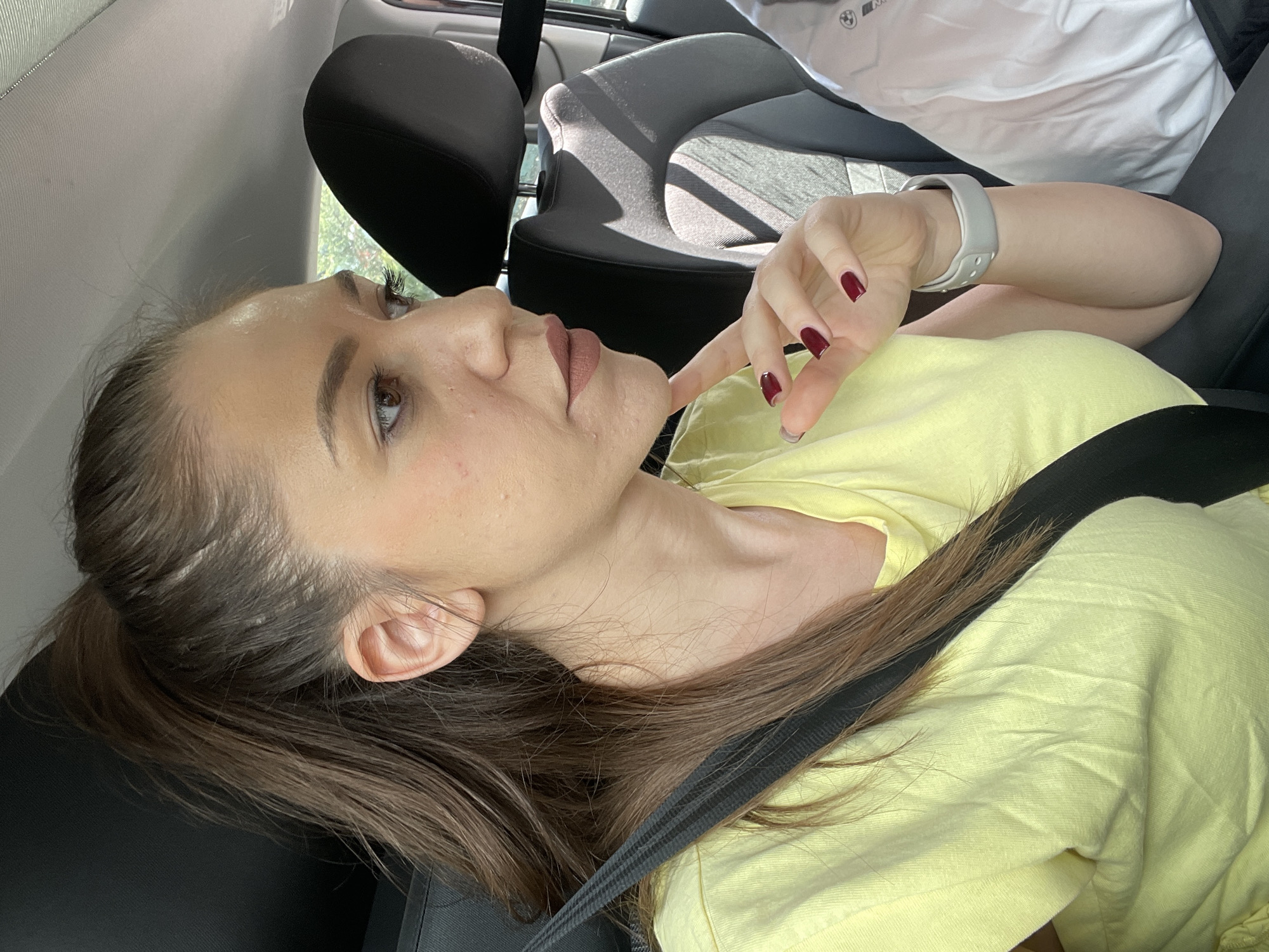 A young woman is depicted in a profile shot from the chest up, seated inside a car. She has long, dark brown hair, which appears to be tied back, and a light yellow t-shirt. Her facial features include well-defined eyebrows, prominent eyelashes, and a subtle, possibly contemplative, expression. Her lips are a muted pink or mauve color. She is wearing a light grey smartwatch on her left wrist, and her fingernails are painted a dark red.
Her right hand is raised, with her index finger gently touching her chin or lower lip. A black seatbelt is visible diagonally across her chest and shoulder, indicating she is buckled in. The interior of the car is visible, featuring dark grey seats with a subtle pattern. Bright sunlight streams into the vehicle from an unseen window, creating distinct shadows on the car seats and interior surfaces, suggesting it is daytime and sunny. In the upper right background, a partial "BMW" logo and text are visible on what appears to be a white garment or a car accessory. The scene takes place in a car, with the context being Sofia, Bulgaria.
