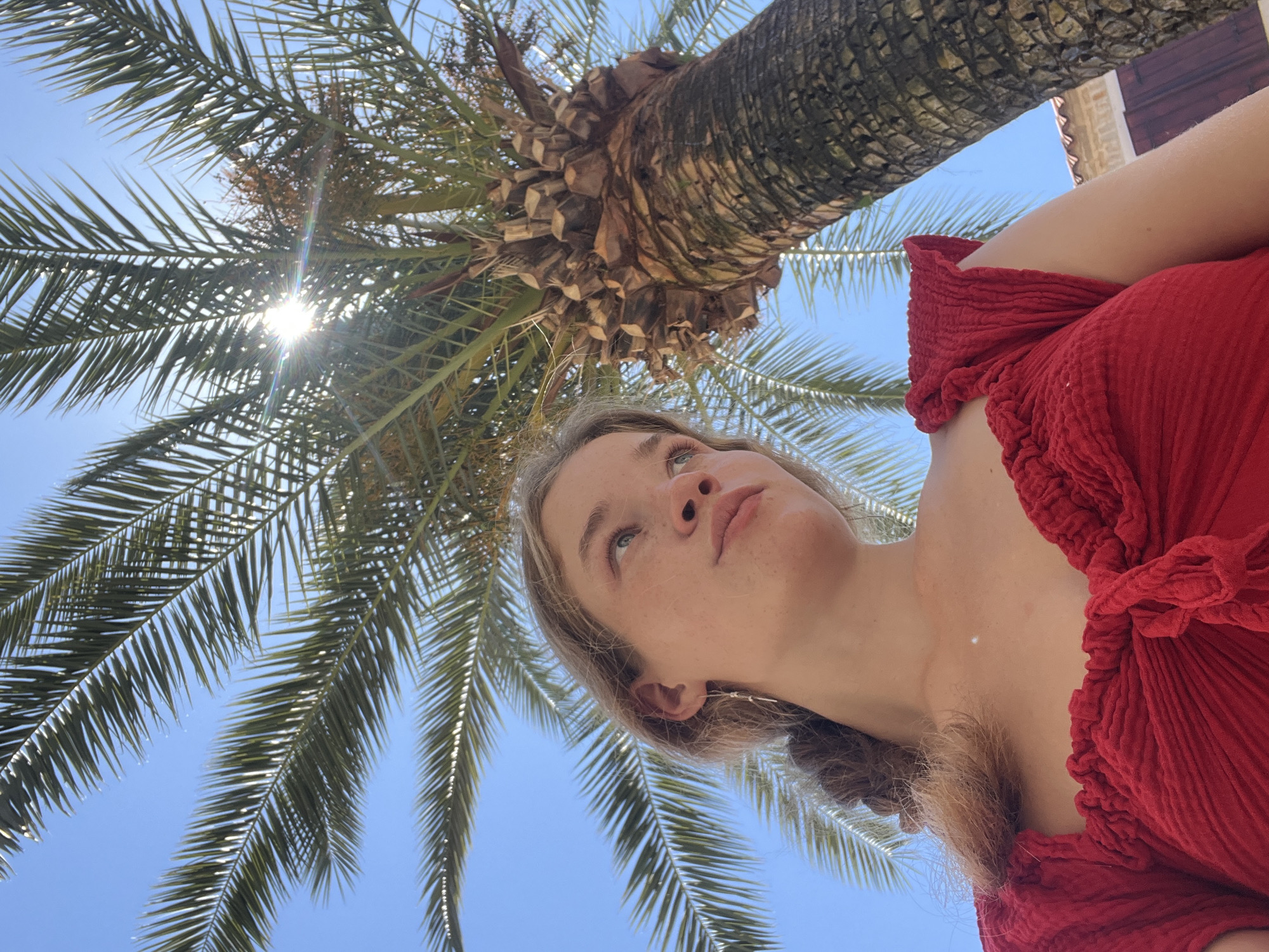The image shows a young woman in a red ruffled top, shot from a low angle looking up. Above her is a palm tree with a clear blue sky visible through its fronds. The sun is shining brightly, creating a lens flare effect through the palm leaves. The background includes a hint of a building with a red-tiled roof, suggesting an outdoor setting. The woman's face is the focal point, as she looks upwards with a neutral expression. The location is likely Budva, Montenegro, based on the combination of palm trees, red-tiled roofs, and a bright, sunny environment.