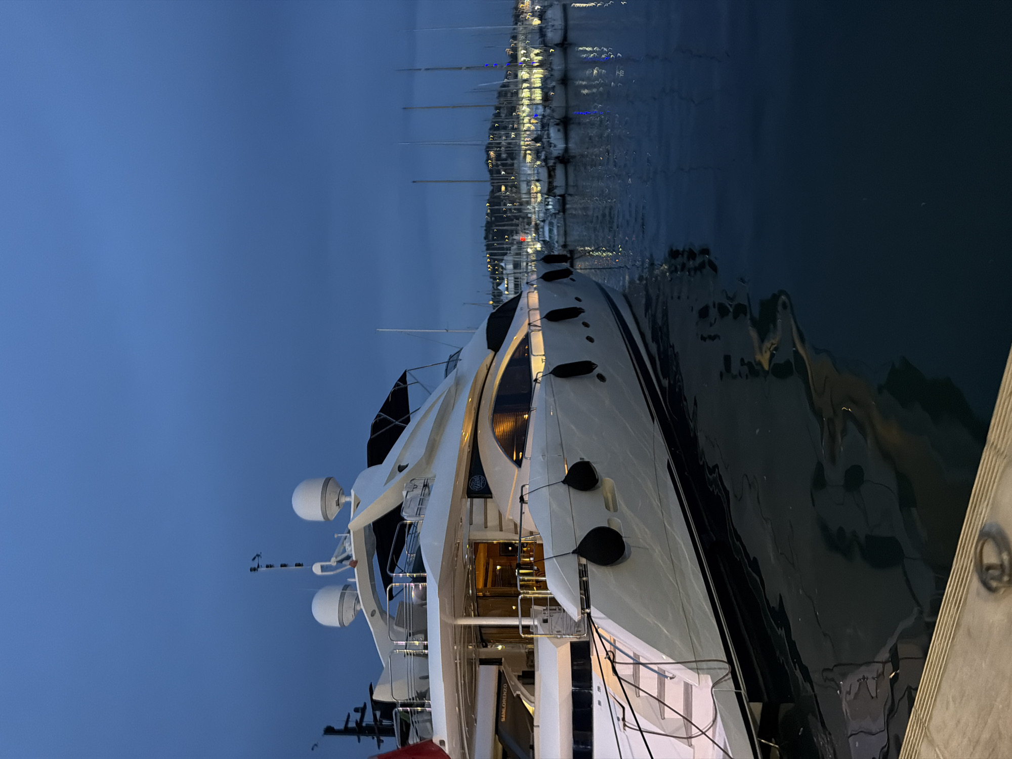 The image captures a luxurious superyacht docked in a marina at dusk or night. The sky is a deep, clear blue, transitioning from lighter blue at the top to darker near the horizon, indicating the evening hours. The main subject is a large, white yacht with black accents, positioned diagonally across the frame, with its bow pointing towards the top left and stern towards the bottom right.
The yacht is brightly lit from within, casting a warm glow from its windows and main deck entrance. Details such as polished railings, white radar domes, and dark fenders are clearly visible on its hull. The water surrounding the yacht is calm and dark, reflecting some of the light from the boat and the distant marina.
In the background, a bustling marina stretches into the distance, lined with numerous other yachts and boats. Their lights illuminate the shore and create sparkling reflections on the water, suggesting a lively port environment. The masts of several sailing boats are silhouetted against the dark sky. To the far right, a sturdy dock or pier structure can be seen, hinting at the yacht's mooring.
The overall atmosphere is serene and opulent, characteristic of a high-end maritime location like Cannes, France. Text visible on the side of the main yacht reads "SANLORENZO," identifying its manufacturer. No people are explicitly visible in the frame, emphasizing the quiet elegance of the scene.