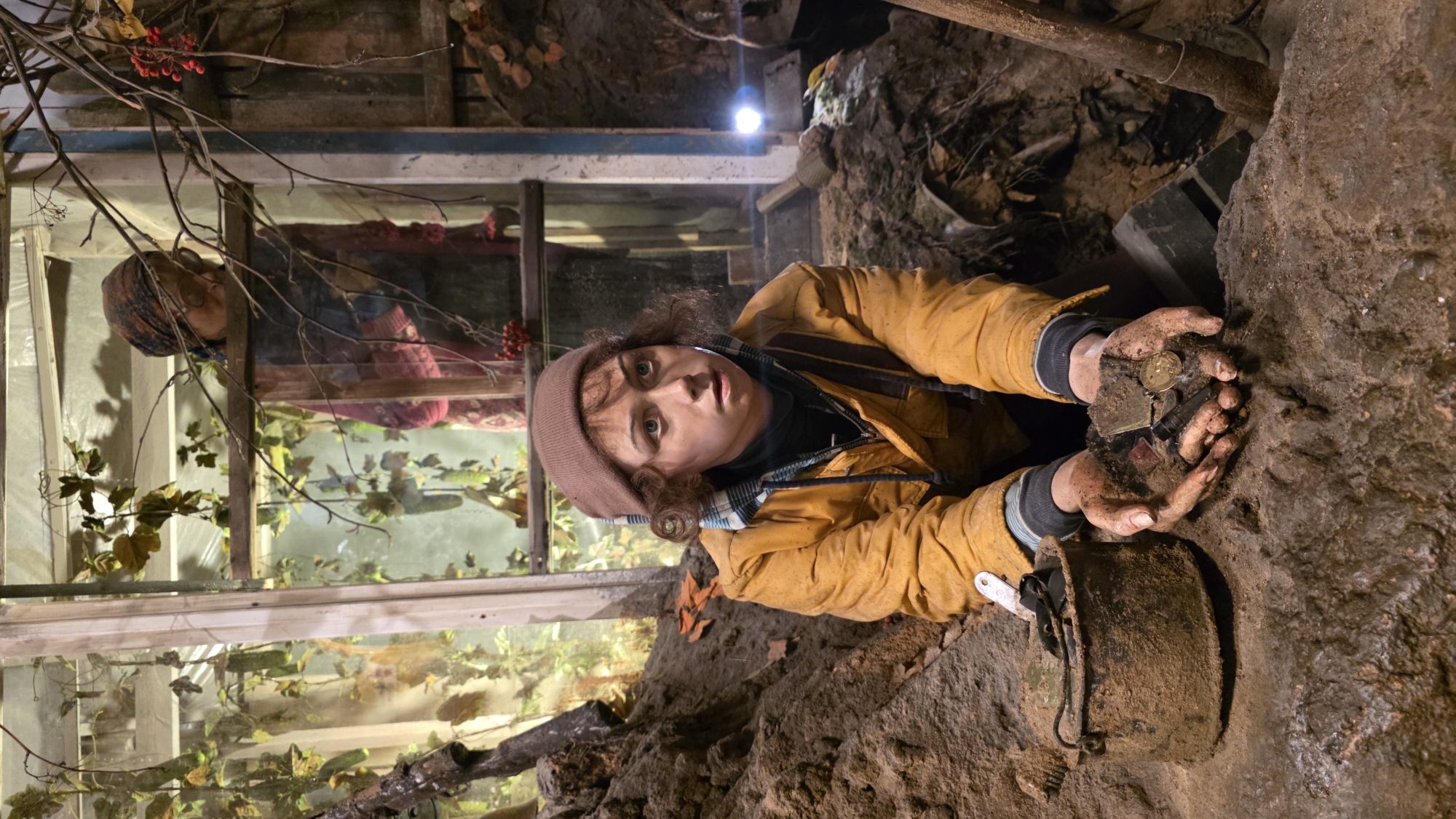 The image shows a woman with curly brown hair, wearing a brown beanie and a mustard-colored jacket, lying on her stomach in what appears to be a narrow tunnel or underground space. Her hands are covered in dirt, and she is holding a collection of small objects, including a coin, a lighter, and other items.
Behind her, a glass-covered structure is visible, possibly a terrarium or window box filled with plants and greenery. Reflected in the glass is the image of an older woman wearing a headscarf, possibly observing the scene. The environment is dark and earthy, suggesting a location like a basement, cave, or underground passage. A lamp provides illumination, casting light onto the scene.