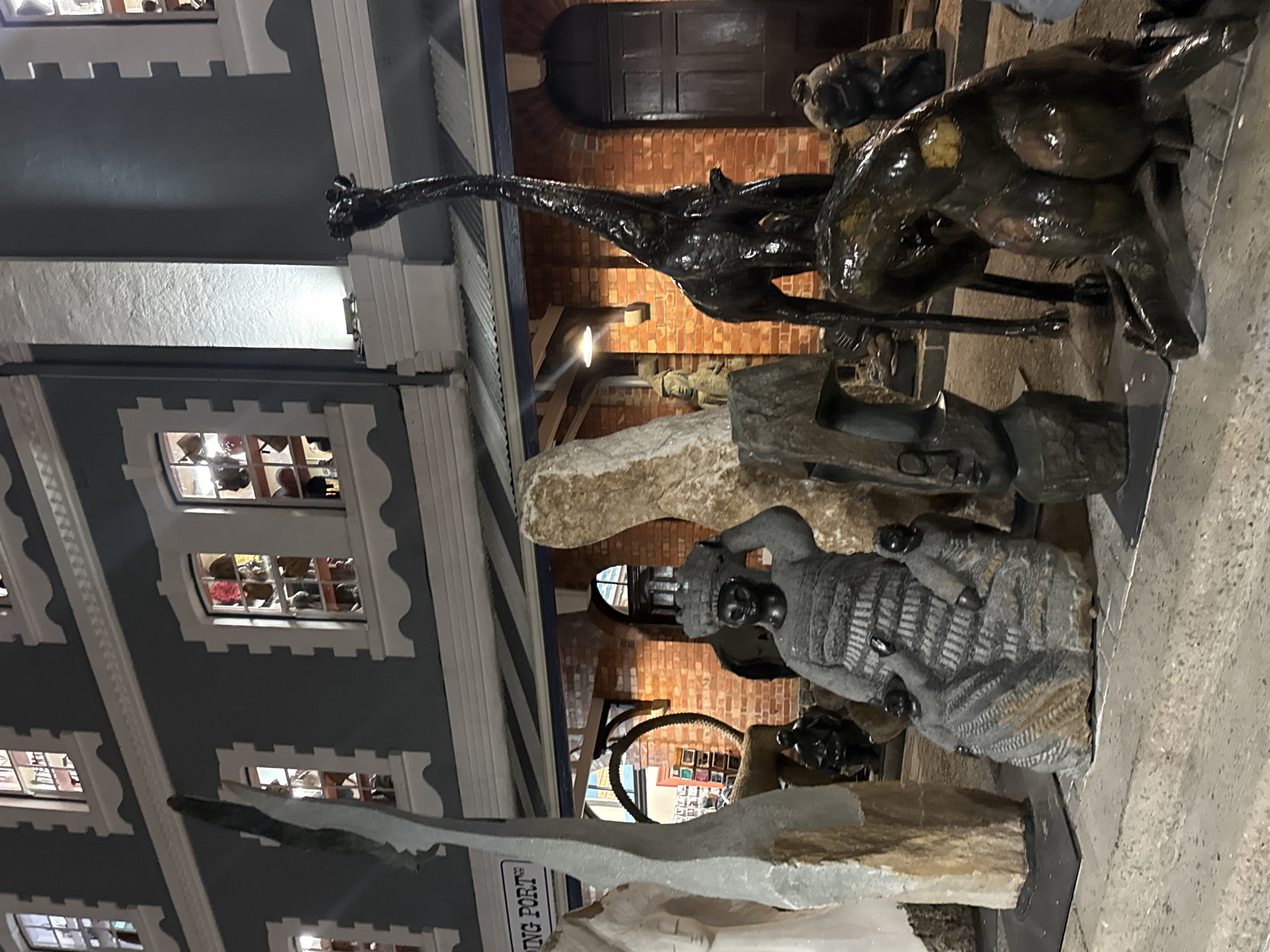 The image shows a collection of sculptures in an outdoor display. Prominent among them are a bronze sculpture of a giraffe and another of a quadruped, possibly a bull or similar animal. Other sculptures include carved stone figures, one resembling a person holding a bowl. The sculptures are arranged in front of a building with a multi-windowed facade, resembling a historic structure or a stylized replica. The location is inferred to be in Cape Town, South Africa, based on identifying the architectural style, signage, and general environment.