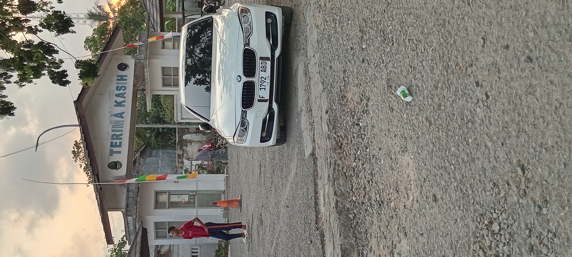 In Kalipucang, Indonesia, a white BMW sedan is parked on a gravelly, cracked asphalt surface in front of a building with an archway. Above the archway, the words "TERIMA KASIH" (Thank You) are visible in blue lettering. A young boy wearing a red t-shirt and dark pants stands to the left of the car, looking to his left. In the background, a person wearing a grey hijab walks away from the camera, and another person on a bicycle is also visible. The environment appears to be outdoors, with trees and foliage in the background. A small white and green object, possibly a discarded cup or container, lies on the ground in the foreground. The sky is cloudy, with a hint of sunset colors.