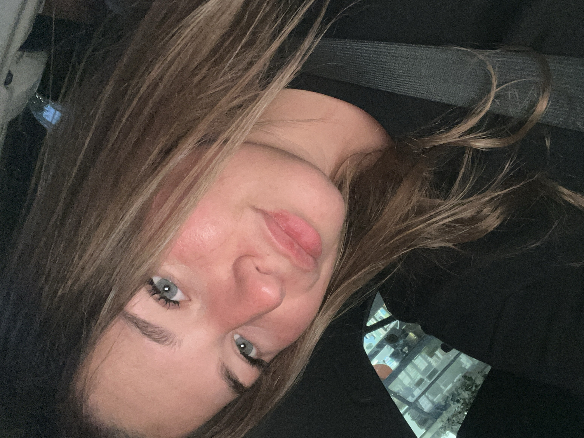 A close-up, rotated portrait captures a young woman with light brown hair and striking blue-gray eyes, looking directly at the camera. Her head is tilted diagonally across the frame, showcasing her fair skin, full pink lips, and a subtle flush on her cheeks. Dark eyelashes frame her eyes, and her eyebrows are well-defined.
She is wearing a dark-colored garment, likely black. A dark gray seatbelt is prominently visible across the upper right portion of the image, indicating she is seated inside a vehicle. In the bottom right corner, a reflective surface, possibly a window or mirror, shows a blurry yet discernible reflection of a building with multiple illuminated windows, suggesting an urban environment. This detail, combined with the subdued lighting, hints at the photo being taken during evening or nighttime in a city such as Budva, Montenegro. Her expression is neutral and composed. There is no visible text.