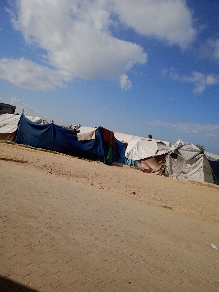 The image displays an outdoor scene in Khan Yunis, Palestinian Territory, under a blue sky with scattered white clouds. The foreground features a light brown paved road constructed from rectangular pavers, sloping gently upwards from left to right. Adjacent to the road, a barren, sandy slope rises, covered with numerous tents and makeshift shelters.
A prominent dark blue tarp, stretched horizontally, forms a large shelter in the mid-left, with a white sheet and other reddish-brown fabrics draped over its exterior. To the right and behind this blue structure, a dense arrangement of white and off-white tents is visible, varying in size and condition. Some tents exhibit standard relief designs, while others appear more rudimentary, constructed from various tarpaulins. One tent in the mid-right features a brown fabric entrance. In the background, atop a more distant white tent, a black water tank is discernible. A partial view of a multi-story, light-colored building with dark rectangular openings is visible in the far left background, situated behind the tents. No individuals are present in the frame. The overall environment appears dry and dusty.