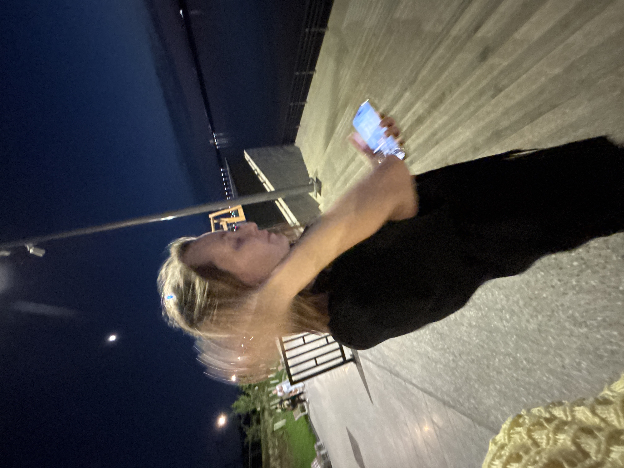 A blurry, low-angle photograph shows a person holding a blue-lit smartphone outdoors at night. The person has light skin and blonde hair. They are wearing a dark shirt. The background includes dark sky, a sliver of moon, a body of water, a street light, green foliage, and a paved walkway. Metal barriers are visible. Based on the visual evidence, the location is consistent with a waterfront area. It is not possible to confirm with certainty that this is Perm, Russia, based solely on this image.