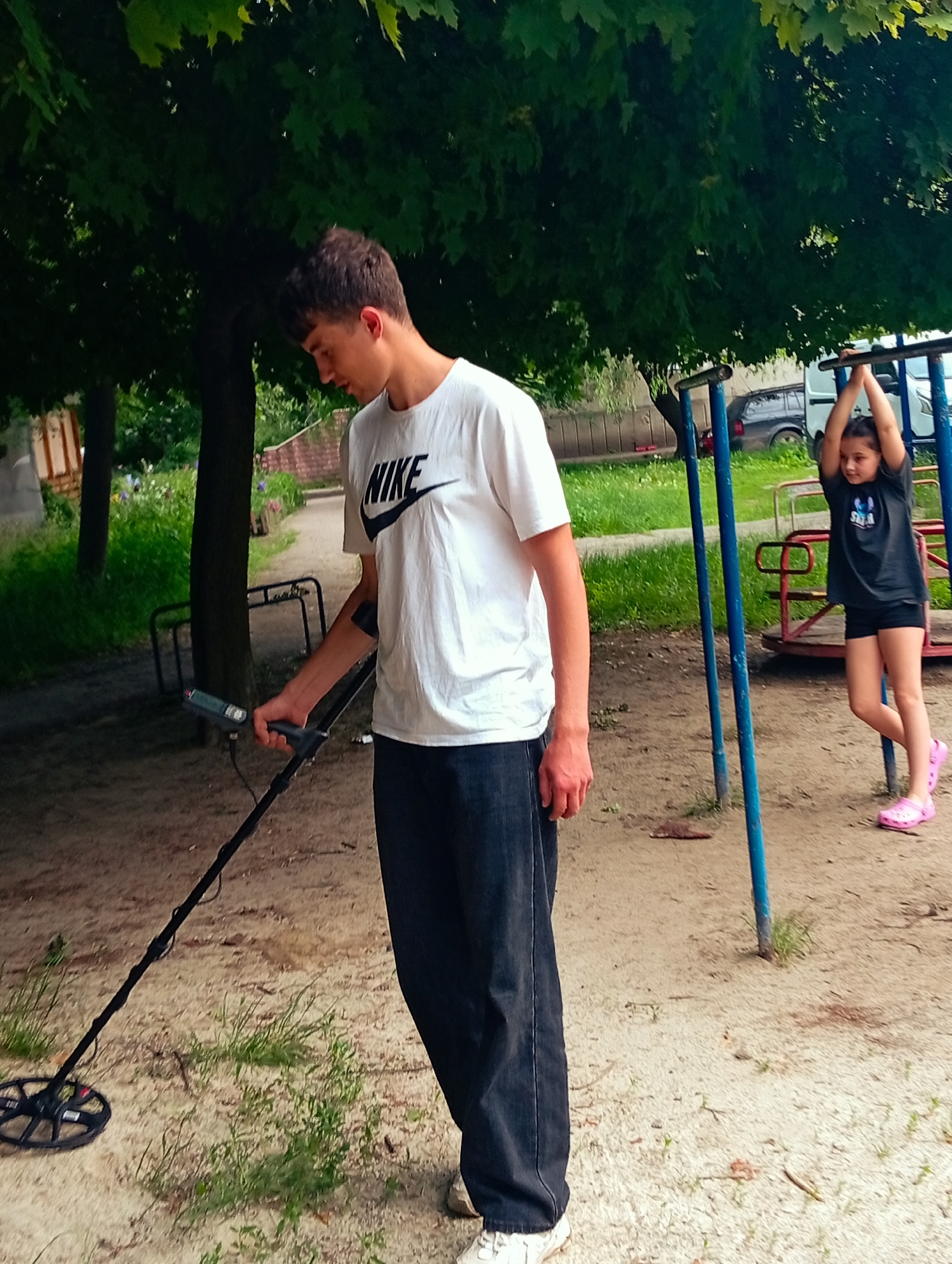 The image captures an outdoor scene featuring a young man and a young girl, likely in a park or a communal green space in Poltava, Ukraine, during daylight hours. The setting is characterized by a dirt ground with patches of grass, shaded by dense green foliage from large trees overhead.
In the foreground, a young man, dressed in a white short-sleeved t-shirt with the black "NIKE" logo on the chest, dark blue jeans, and white sneakers, is intently operating a black metal detector. He holds the device with his right hand, guiding its circular search coil close to the ground, suggesting he is searching for hidden objects. His head is slightly bowed, indicating focus on his task.
Further back on the right side of the frame, a young girl is engaged in play on playground equipment. She is seen hanging from blue horizontal bars, with her arms extended upwards. She wears a dark t-shirt with some faint white text, black shorts, and bright pink footwear, resembling Crocs. Behind her, part of a red merry-go-round is visible. In the background, a dark car and a white van are parked, hinting at a nearby road or residential area. The lighting suggests an overcast or partly cloudy day, providing soft, diffused light without harsh shadows.