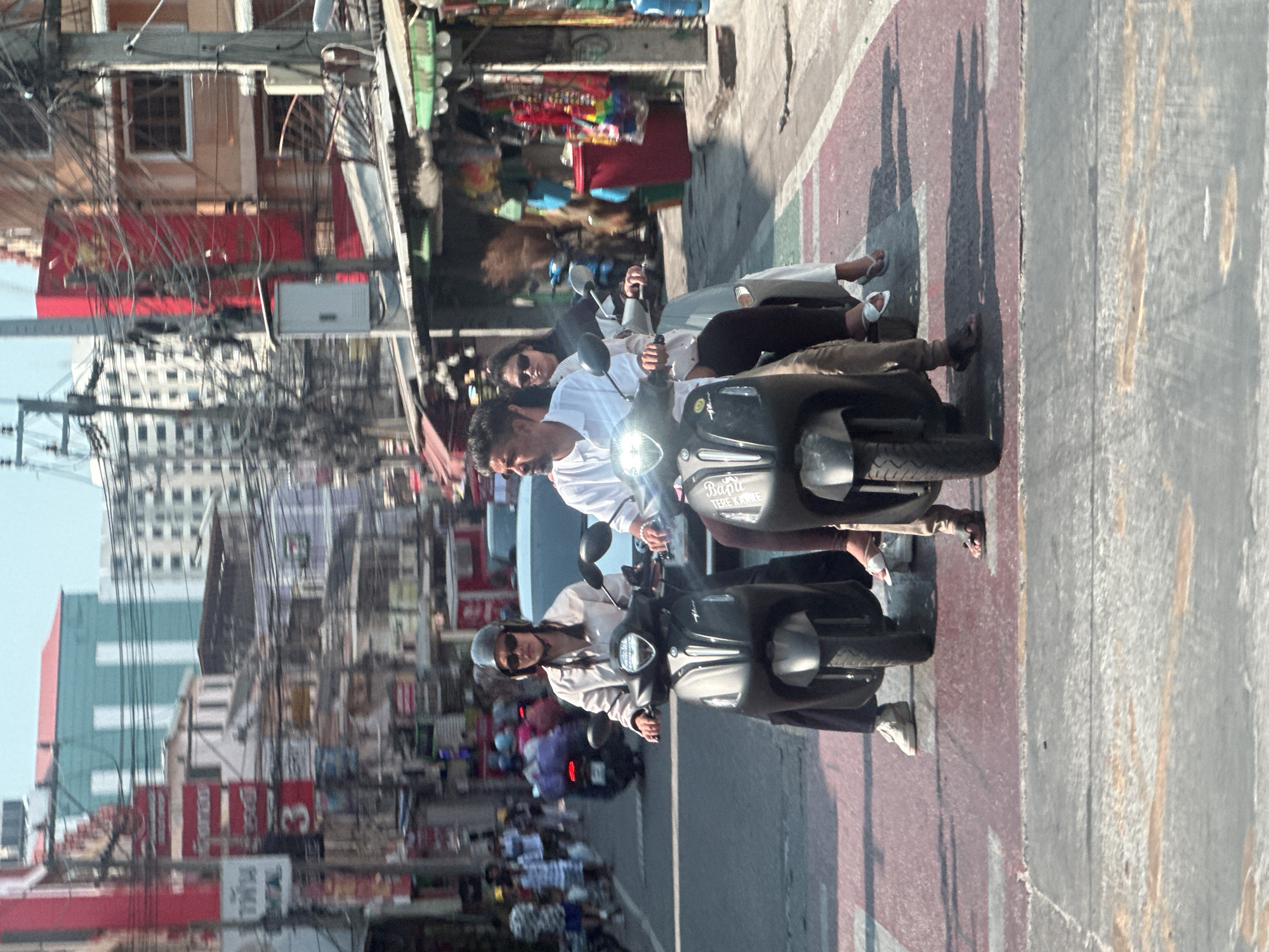 Here's a detailed description of the image:
The image captures a street scene in Pattaya City, Thailand. Two motorbikes are prominently featured, moving along a road marked with red and white stripes. The lead motorbike is ridden by a woman wearing a helmet and sunglasses, while the second motorbike carries a man in a white shirt and another passenger behind him.
The backdrop reveals a typical urban scene. Buildings of varying heights and architectural styles line the street. Overhead, a dense network of electrical wires creates a complex web against the sky. Pedestrians can be seen on the sidewalks, and other motorbikes populate the road. Shop fronts are visible, displaying merchandise. The overall setting suggests a bustling commercial area within the city.