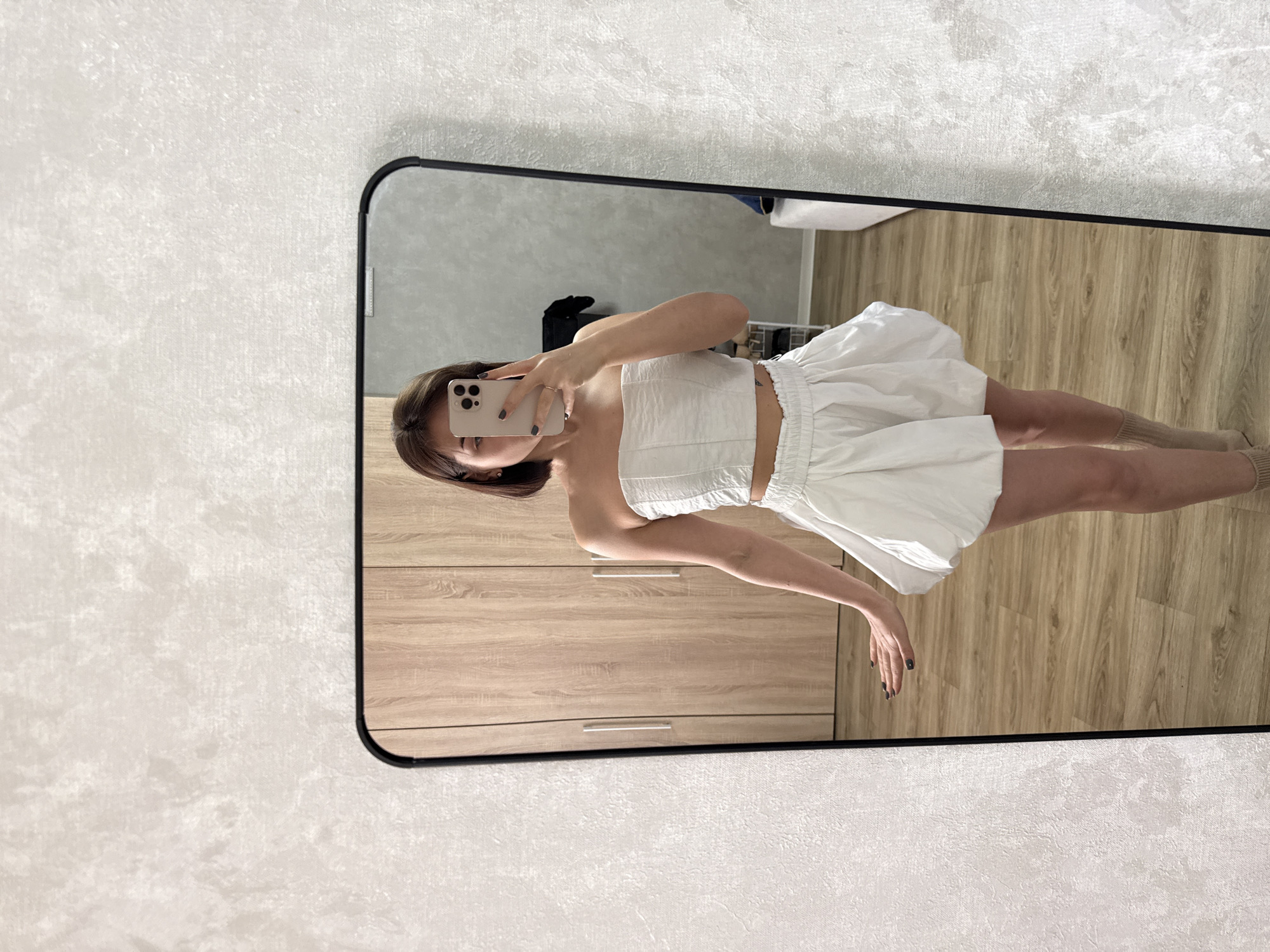 A woman is taking a selfie in a mirror. She's wearing a white strapless top and matching white shorts. She is in a room with light wood floors and light wood cabinets, with a light-colored patterned wall partially visible. The location is Лобаново, Russia.