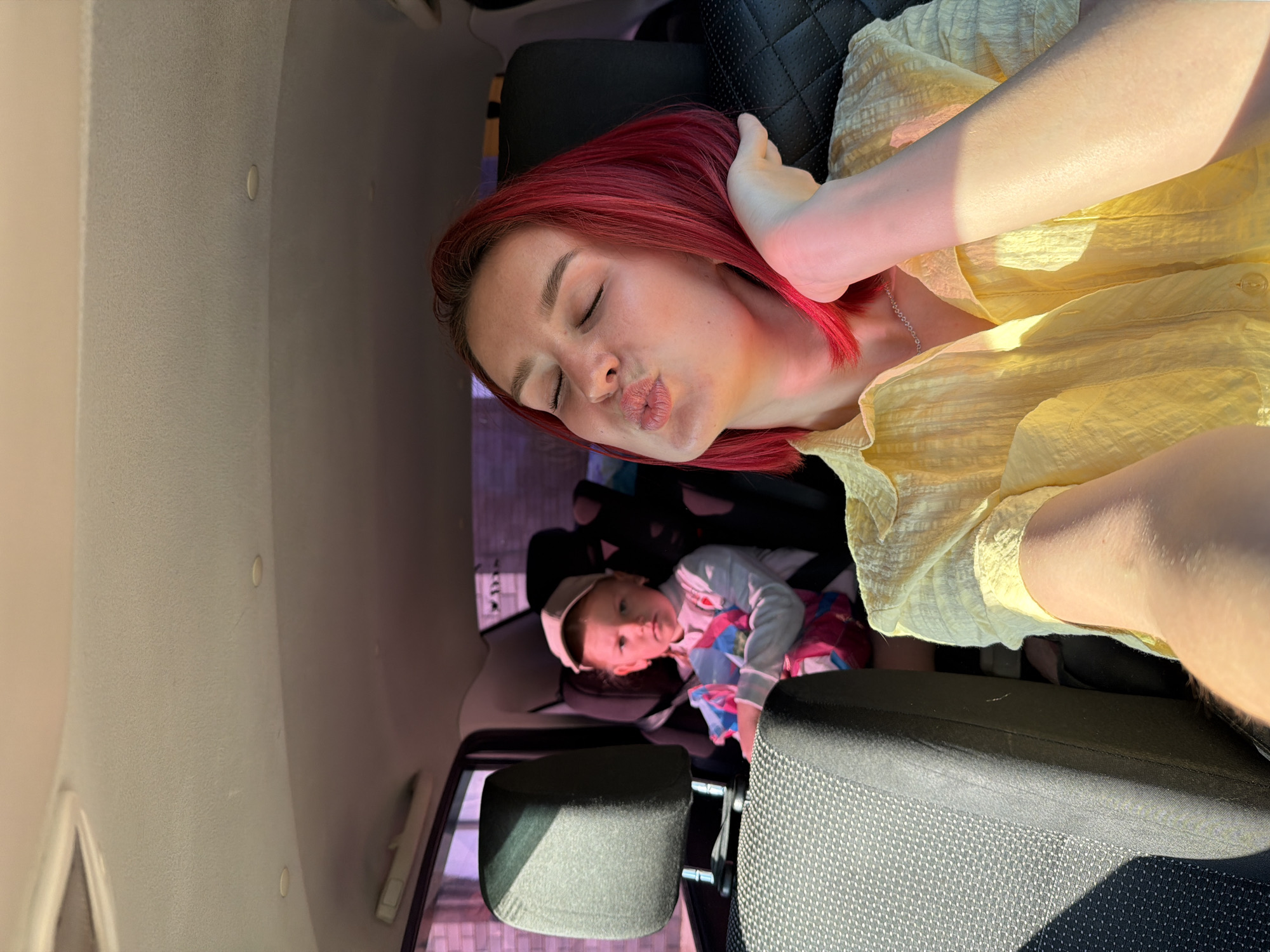 A woman with red hair, wearing a yellow shirt and silver necklace, is seated in a car and making a kissing face. Behind her is a child in a car seat. The interior of the vehicle is visible, including the seat material and car windows. The location is Chaikovsky, Russia.