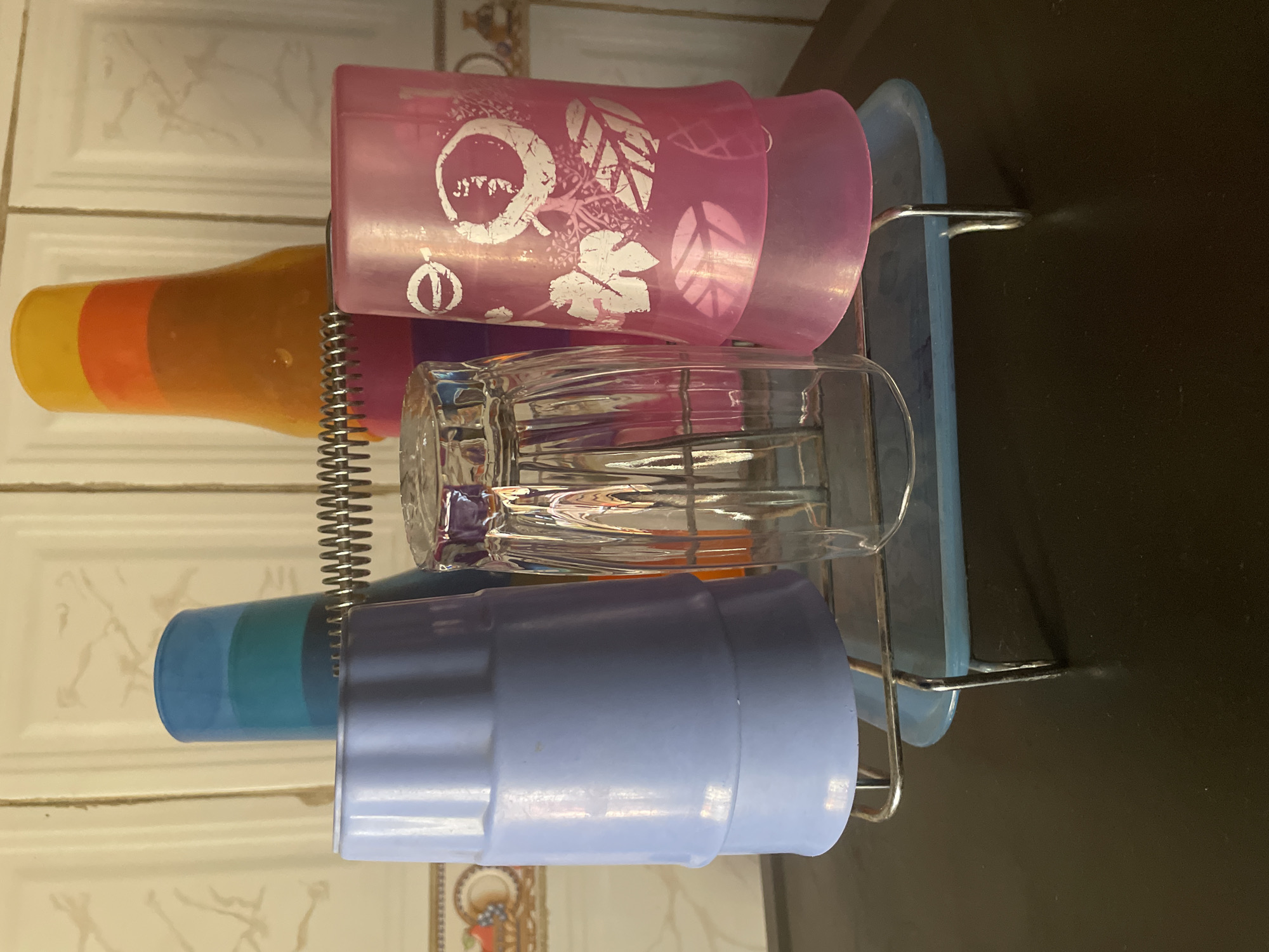 A rack holds four cups and two bottles in front of a tiled wall. The rack is made of silver metal and has a blue plastic base. The cups are stacked on the rack. From left to right, there is a blue cup, a clear glass cup, and a pink cup with a leaf design. Behind these cups are two bottles. The bottle on the left is blue and green. The bottle on the right is orange, yellow, and red. The tiled wall is cream colored with a faint pattern. This scene is located in the city of Kakau, Nigeria.