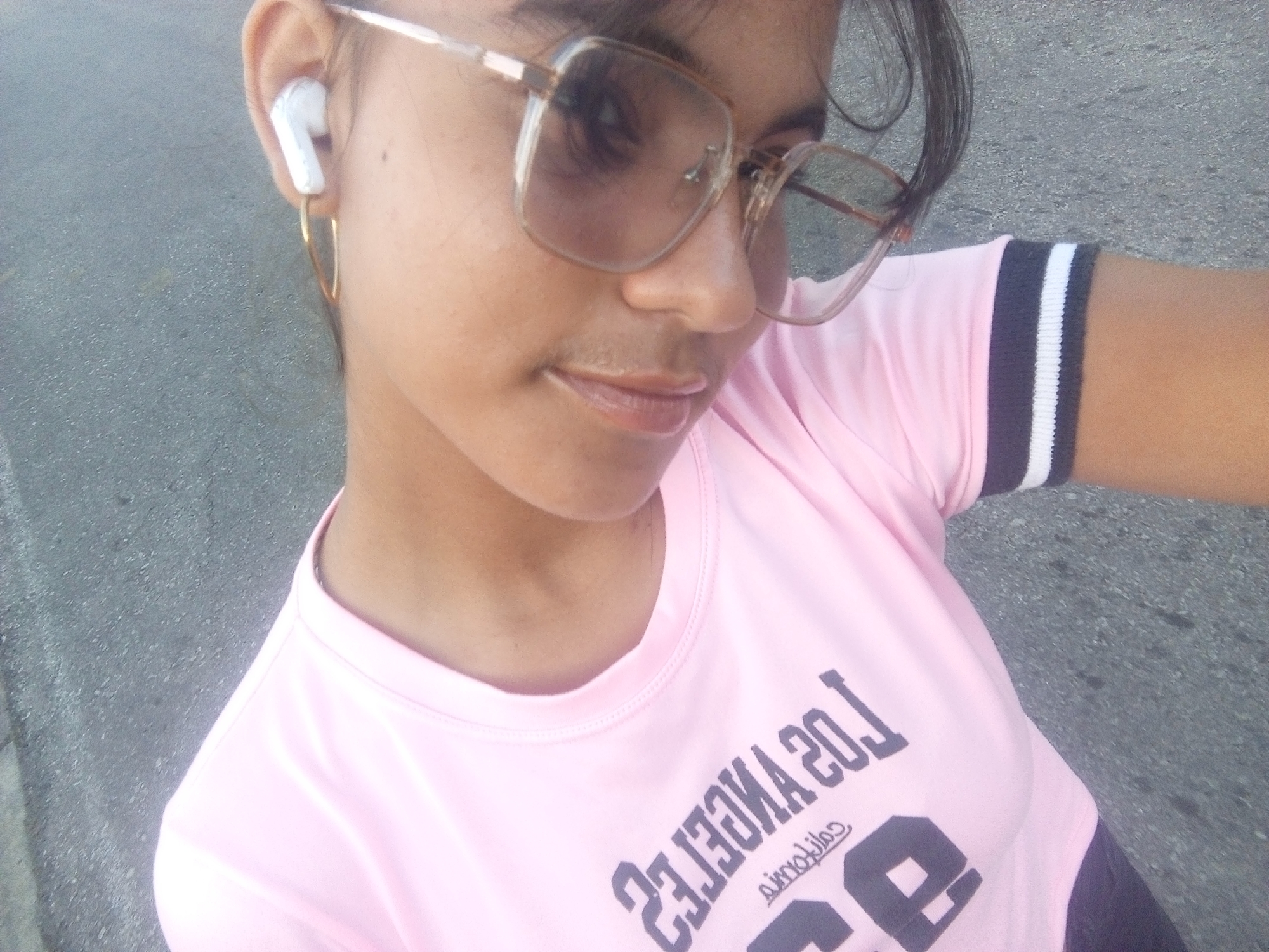 A woman is outdoors in an urban setting. She is wearing sunglasses, a pink t-shirt with the words "Los Angeles 20" printed on it, and white wireless earbuds in her left ear. She also has a hoop earring in her left ear. The surface below her appears to be asphalt or concrete.