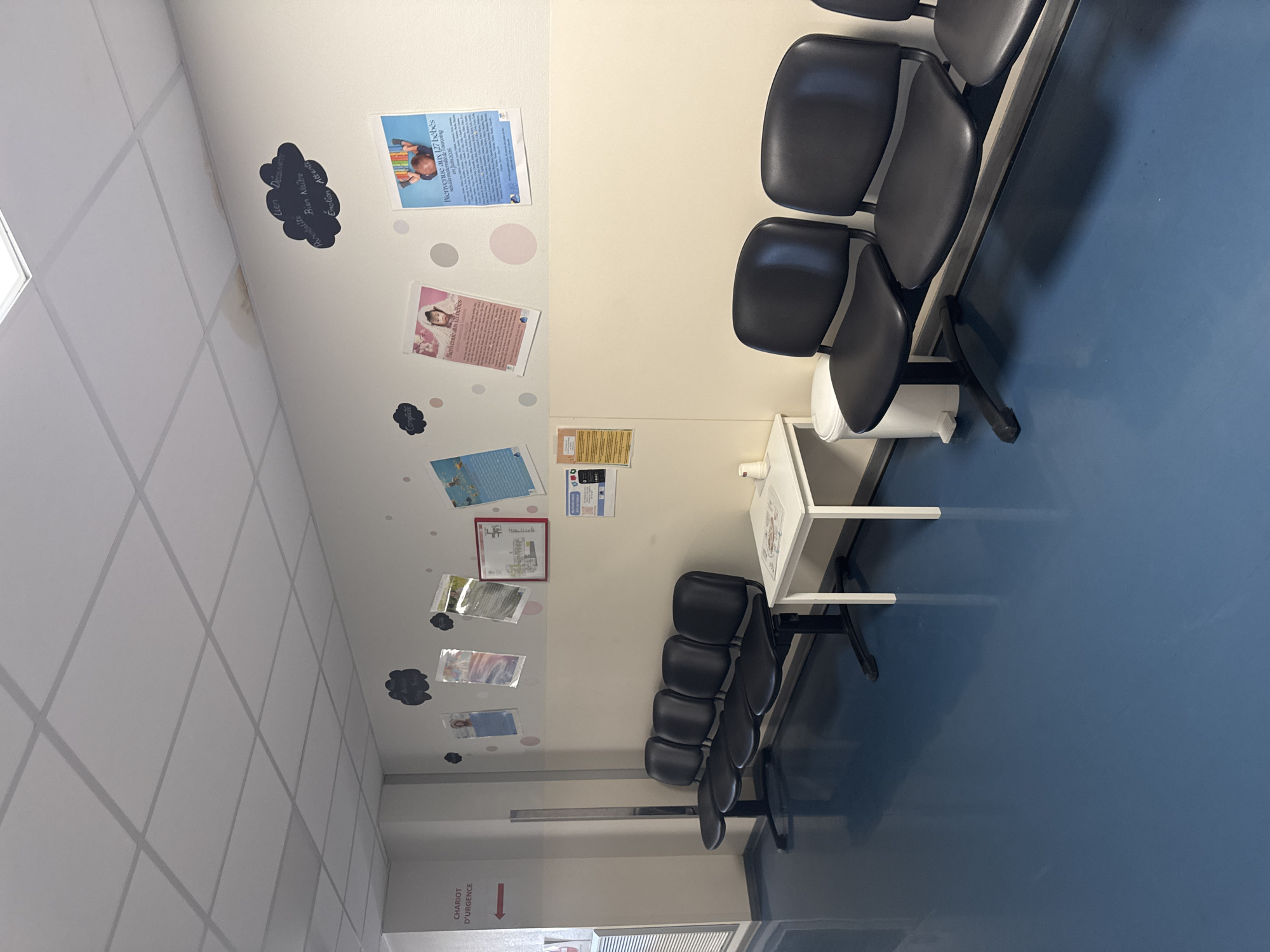 This image depicts a waiting room with black chairs and a blue floor. On the wall, there are several posters and drawings, including a cloud-shaped drawing with handwritten text. A small white table with a trash can is positioned in front of some of the chairs. A sign on the left wall reads "CHARIOT D'URGENCE" with an arrow pointing left, indicating an emergency cart. The scene appears to be in a medical facility or hospital in Tourcoing, France, suggested by the presence of posters related to babies and the mention of "127 bébés accueillis au sein de Tourcoing".