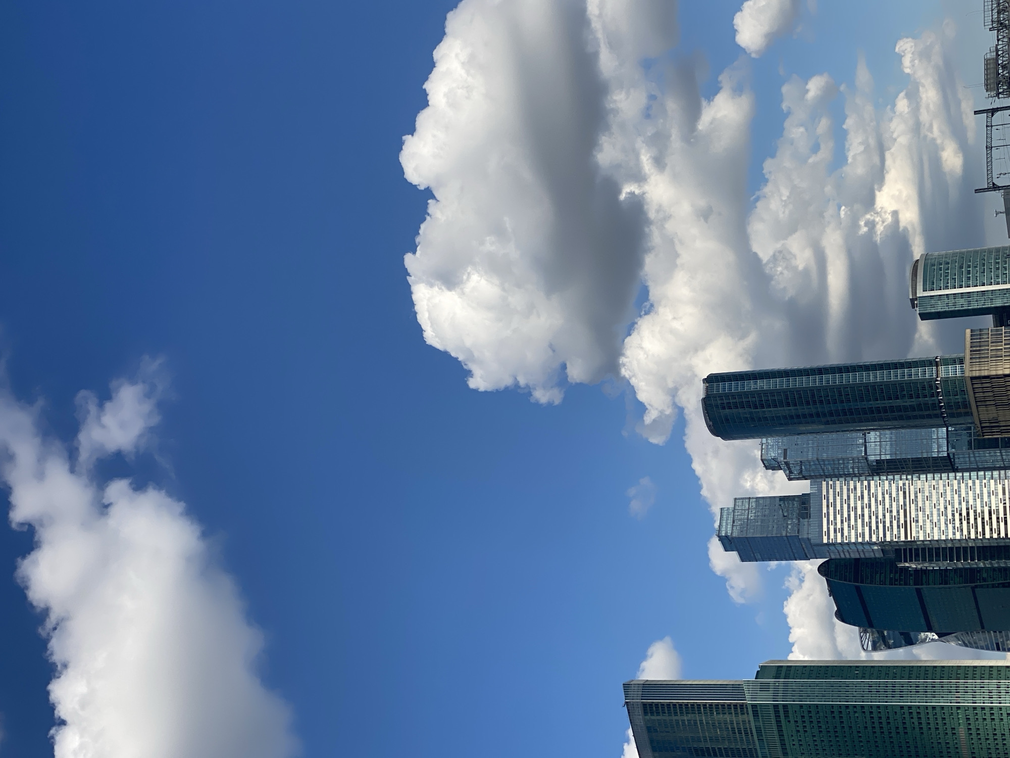 In an urban skyline under a bright blue sky dotted with fluffy white clouds, a cluster of modern skyscrapers pierces the horizon. The buildings exhibit a variety of architectural styles, from sleek, glass-encased towers to structures with more angular and curved designs. The skyscrapers are predominantly dark blue, green, and silver, reflecting the sunlight. The environment appears to be an open area within a city, with a clear view of the sky and distant structures. There are no people visible in the image.