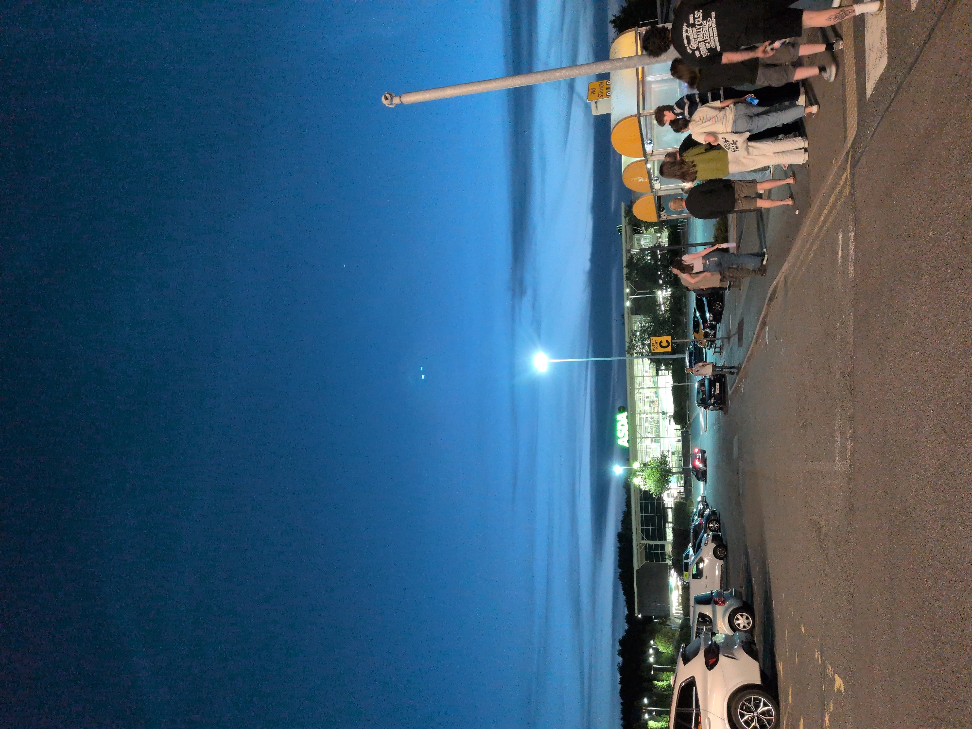 The image depicts a nighttime scene at an ASDA supermarket in Telford, United Kingdom, possibly near a parking lot. The sky is a deep blue, with some faint clouds. In the background, the ASDA store is illuminated, with its logo prominently displayed. Several cars are parked in the lot, and a few streetlights are visible. In the foreground, a group of people are standing near a covered structure with yellow awnings, which appears to be a pay station. Some individuals are walking, while others are gathered in conversation. The overall atmosphere suggests a busy evening at the supermarket.